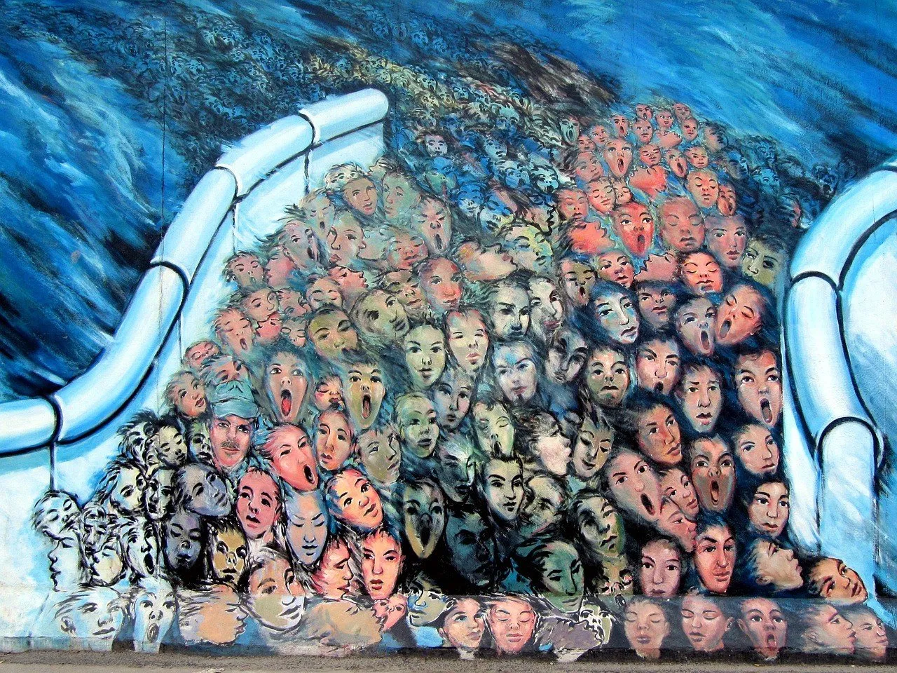 Detalhe de mural da East Side Gallery com muitos rostos entre formas brancas e fundo azul.