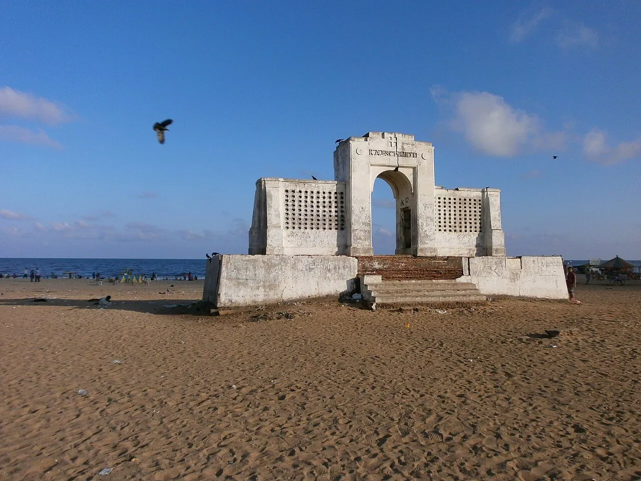 Memorial branco Kaj Schmidt na areia de Elliot’s Beach, com mar azul, aves e visitantes distantes.