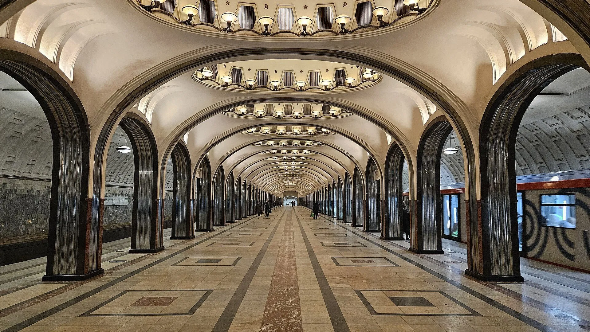 Interior da estação Mayakovskaya de Moscou, com arcos, nichos iluminados no teto e trem à direita.
