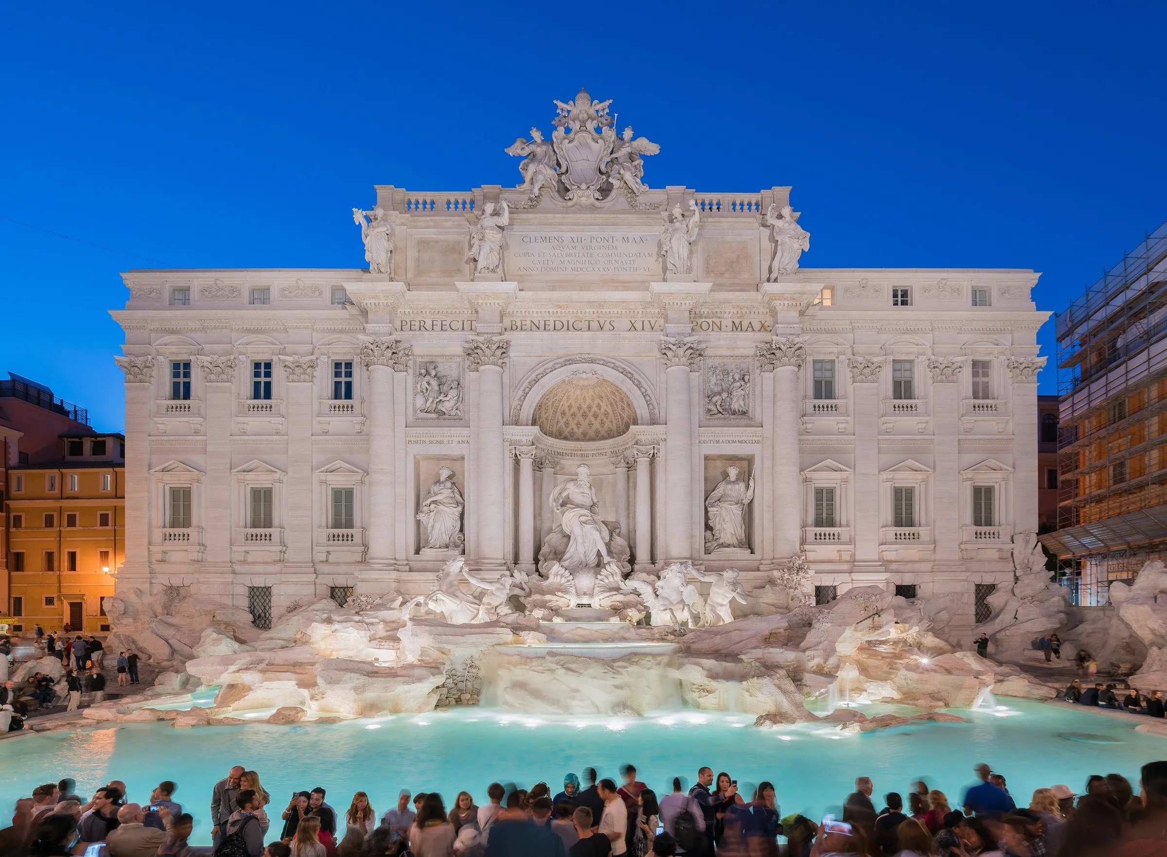 Fachada da Fontana di Trevi iluminada ao entardecer, com a bacia de água turquesa e visitantes na beira.