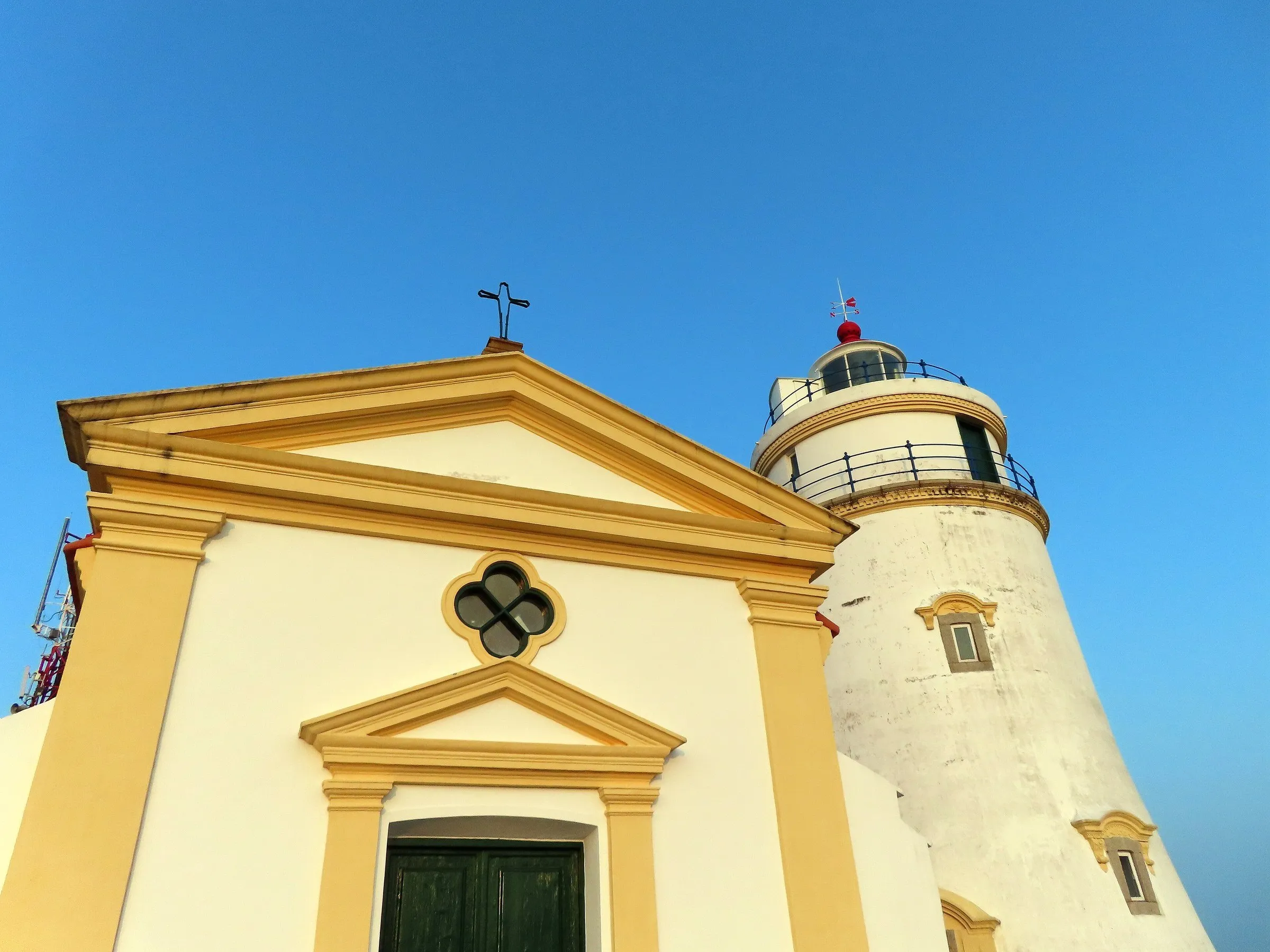 Capela de fachada branca e amarela ao lado do Farol da Guia, torre branca com grade azul, contra um céu azul limpo.