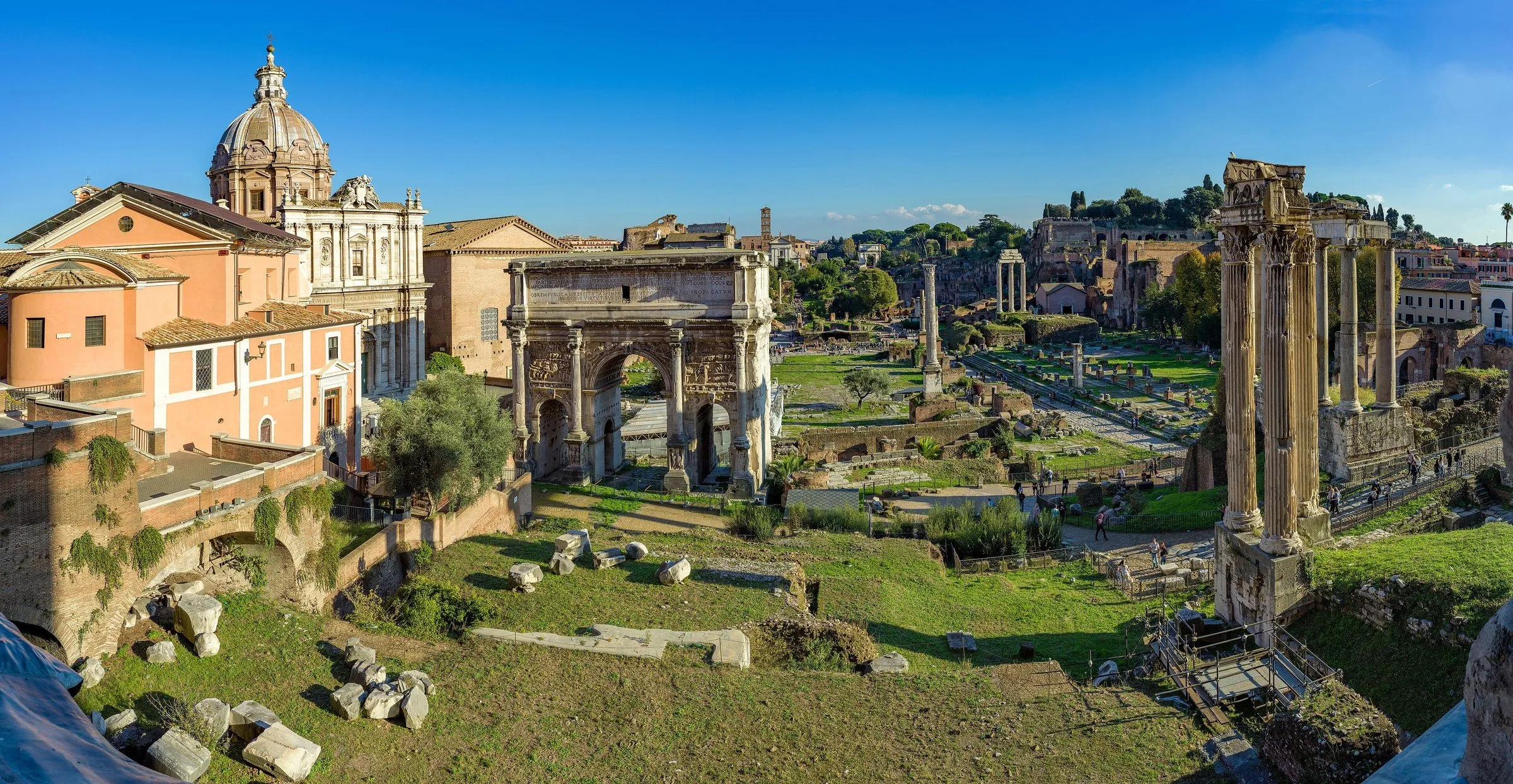 Vista elevada do Fórum Romano, com colunas do Templo de Castor e Pólux, o Arco de Sétimo Severo e igrejas ao fundo.