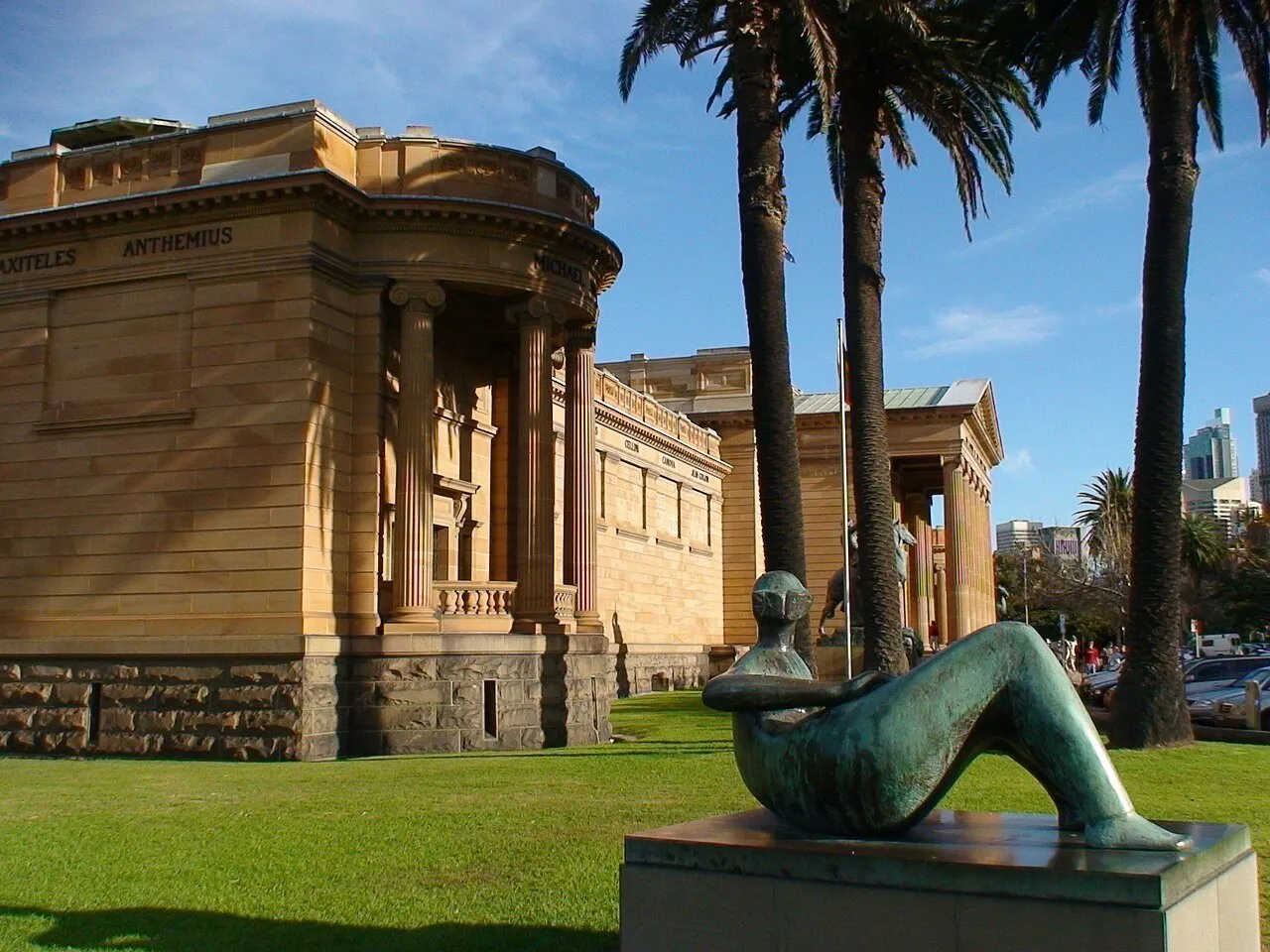 Exterior de pedra da Galeria de Arte de Nova Gales do Sul, com colunas, palmeiras e escultura reclinada.