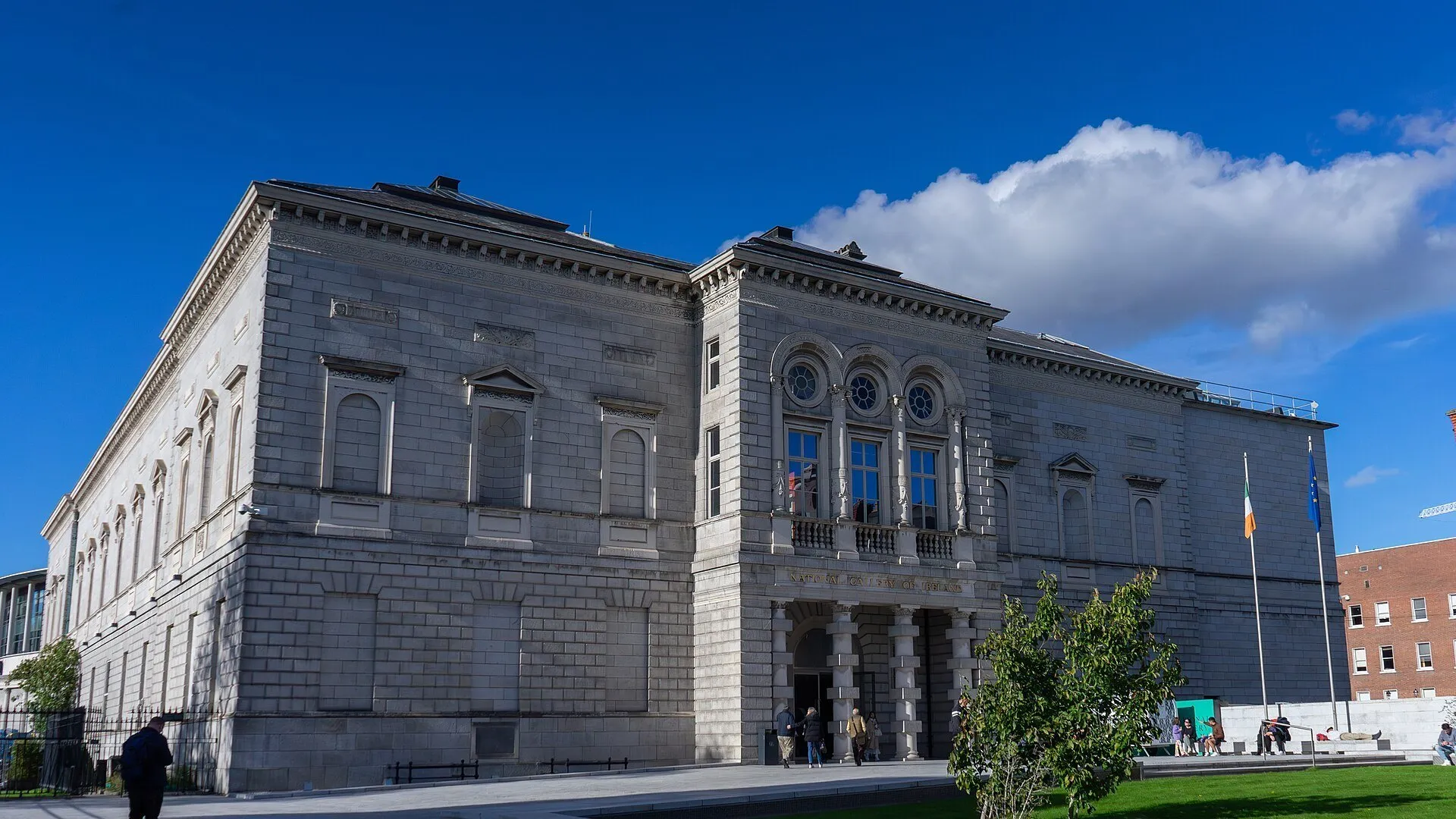 Fachada de pedra da Galeria Nacional da Irlanda com janelas circulares, bandeiras e pessoas diante da entrada.
