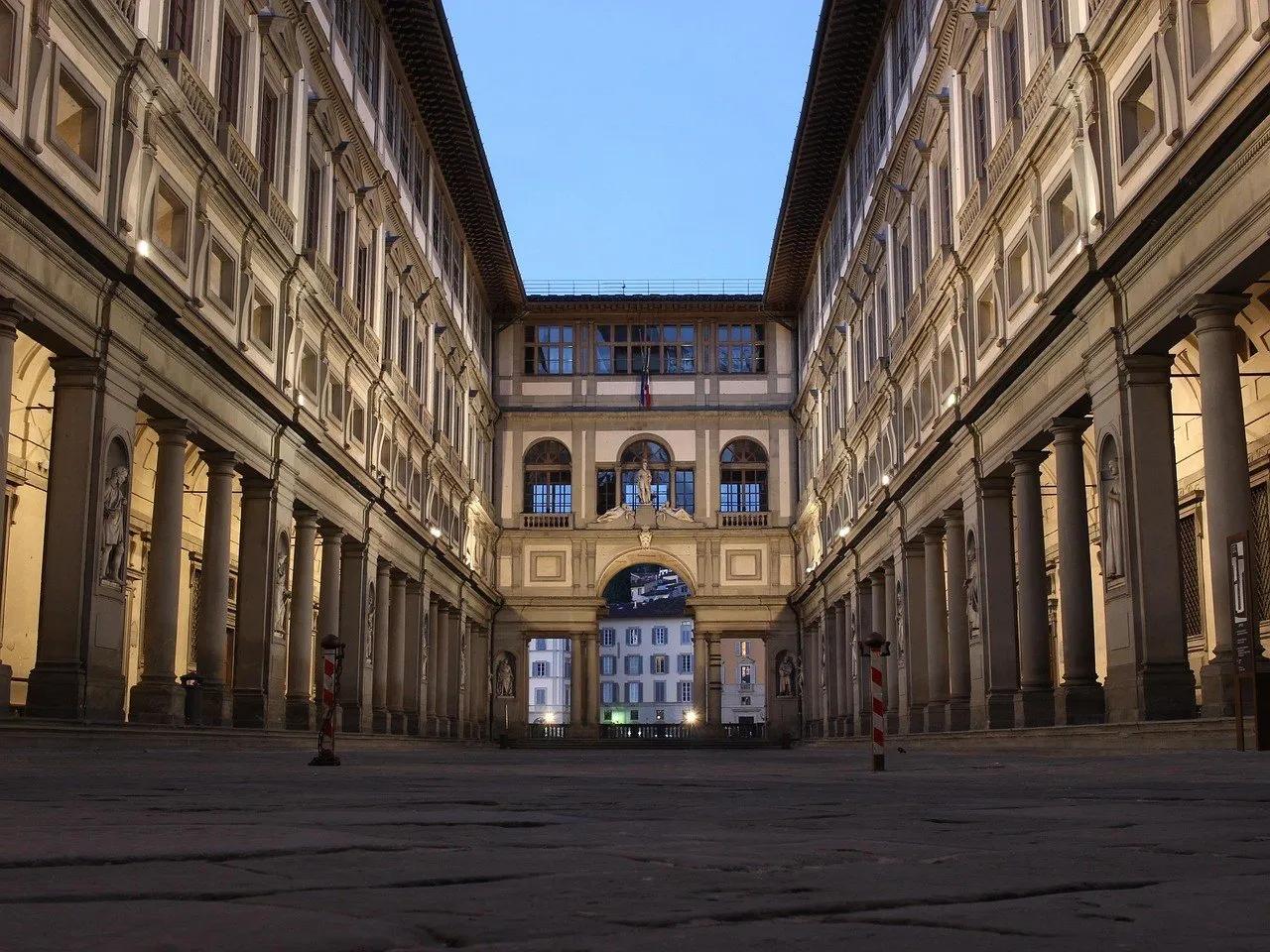 Pátio dos Uffizi com alas de colunas simétricas, iluminação acesa e céu azul do crepúsculo.