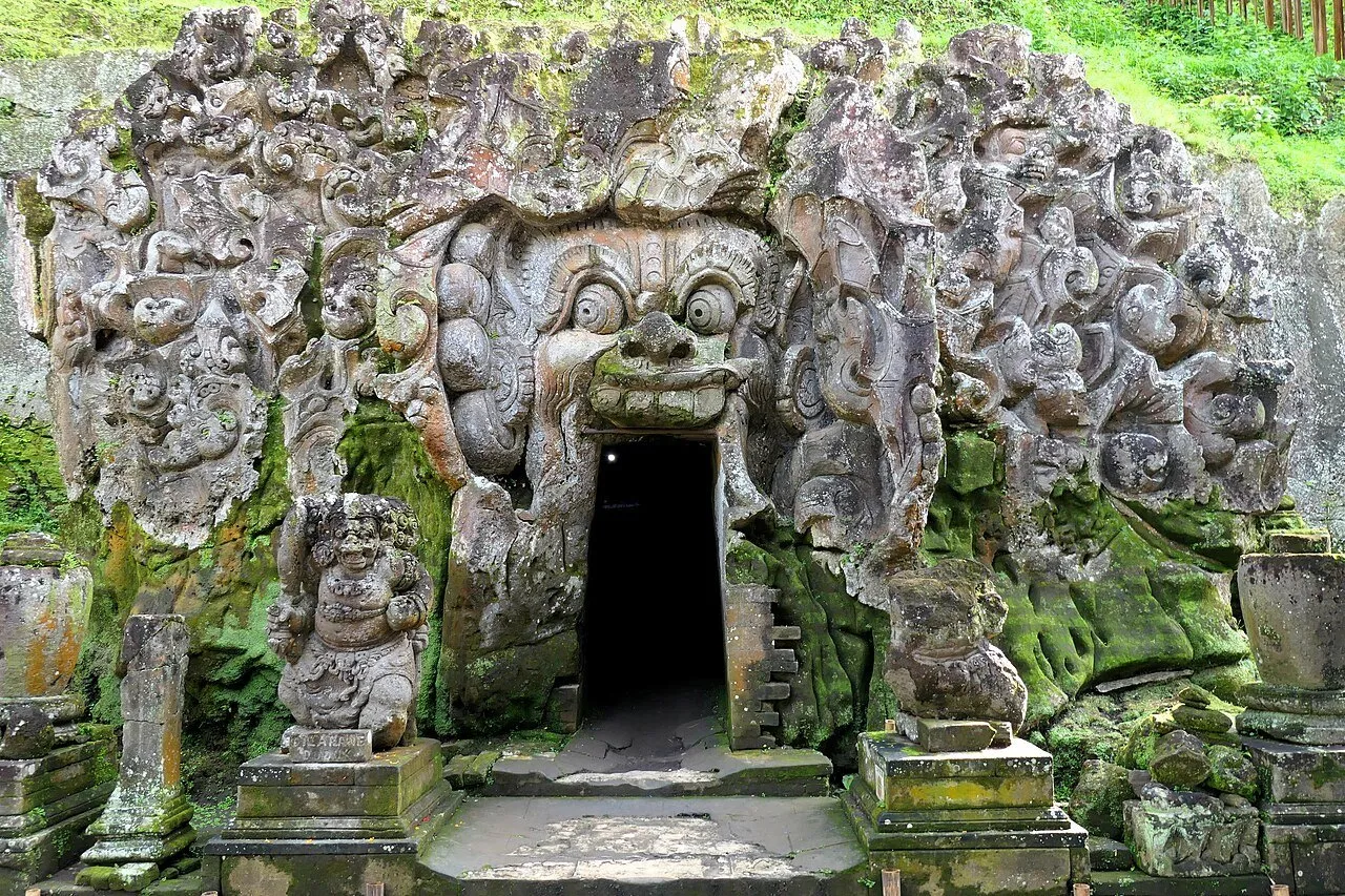 Entrada escura da caverna de Goa Gajah emoldurada por rosto e relevos esculpidos na pedra, com musgo.