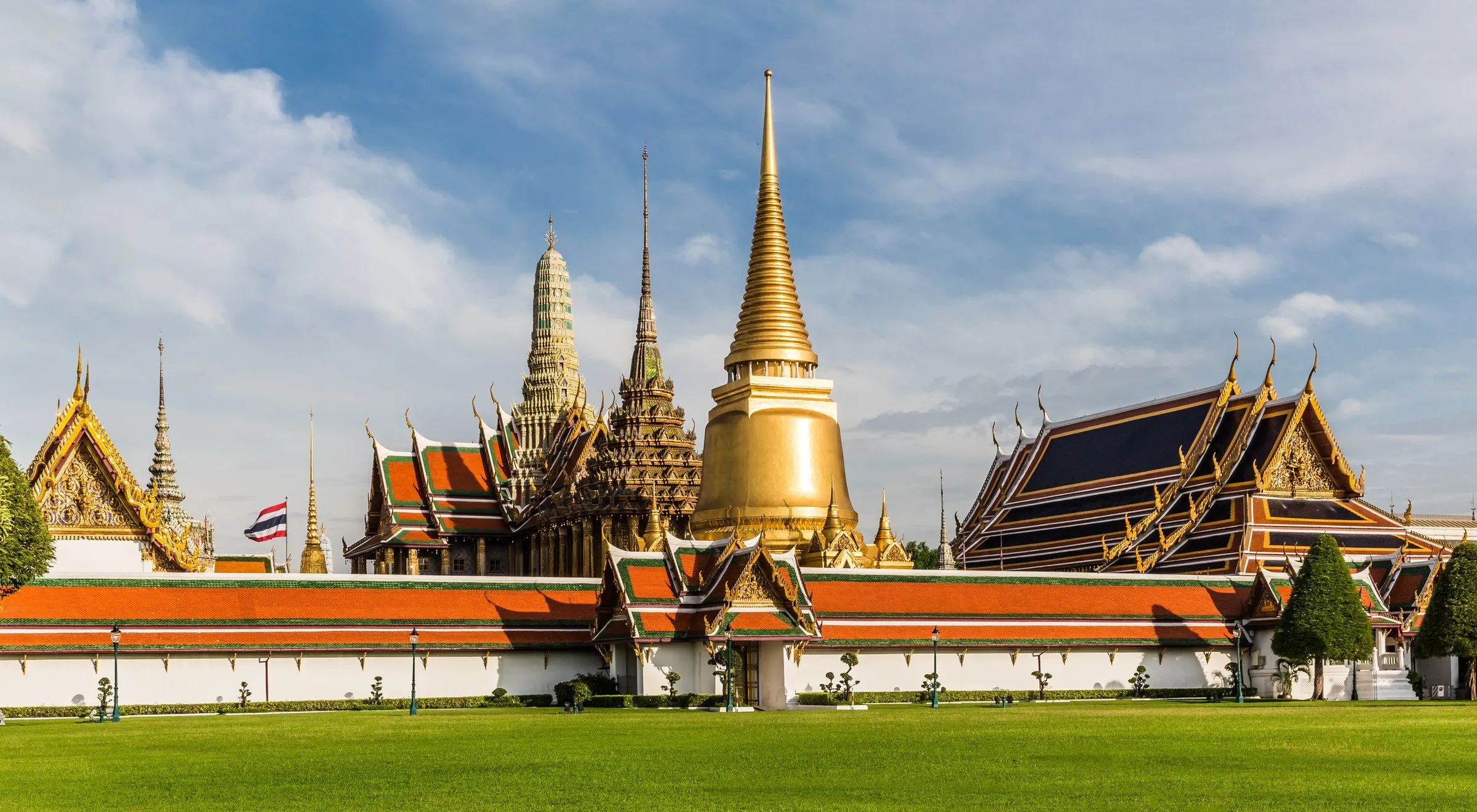 Telhados laranja e pináculos dourados do Grande Palácio, em Bangkok, atrás de uma muralha branca e gramado, sob céu azul.