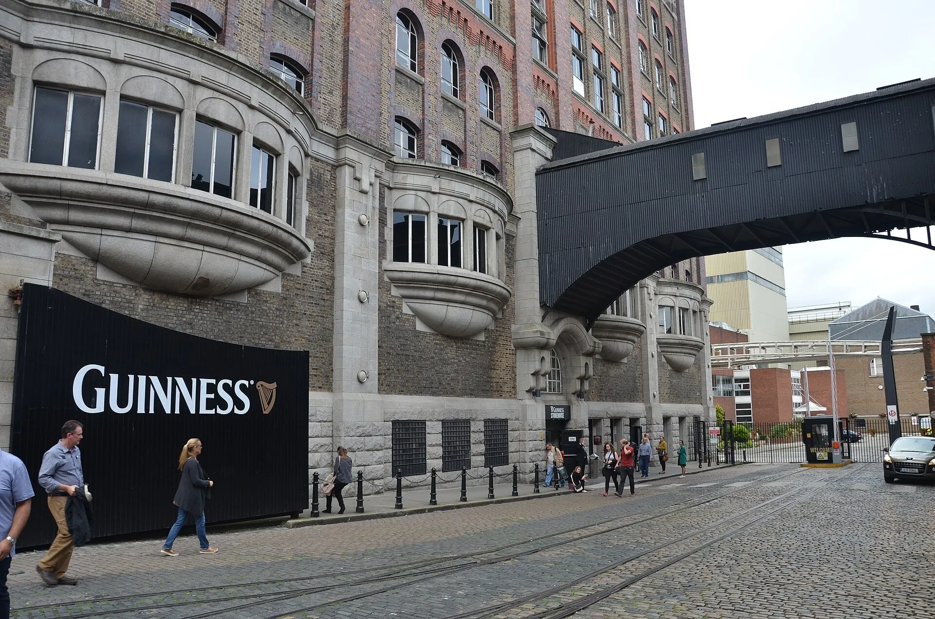 Fachada do Guinness Storehouse com janelas curvas, passarela elevada, visitantes e rua de pedra.