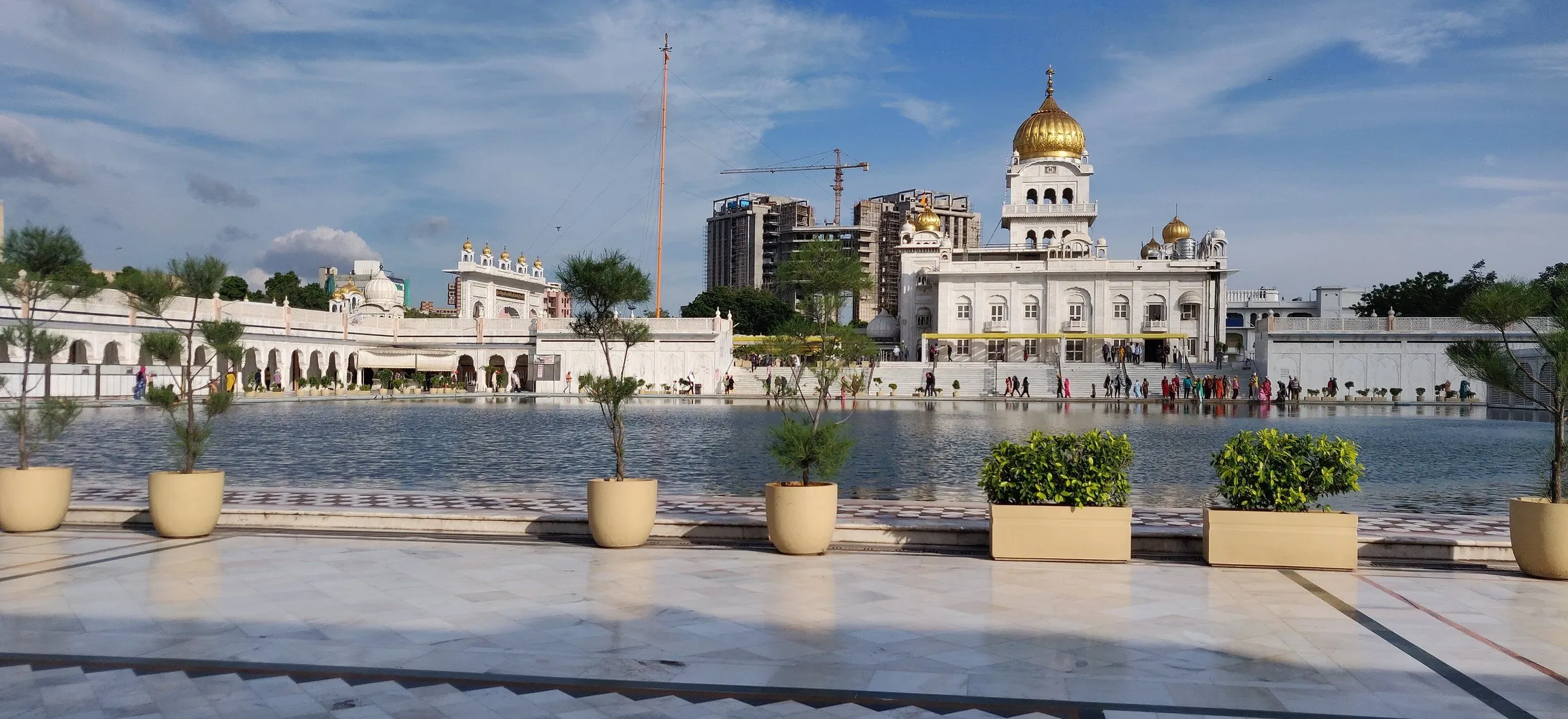 Cúpula dourada do Gurdwara Bangla Sahib atrás do lago sagrado, com vasos de plantas no piso de mármore em primeiro plano.