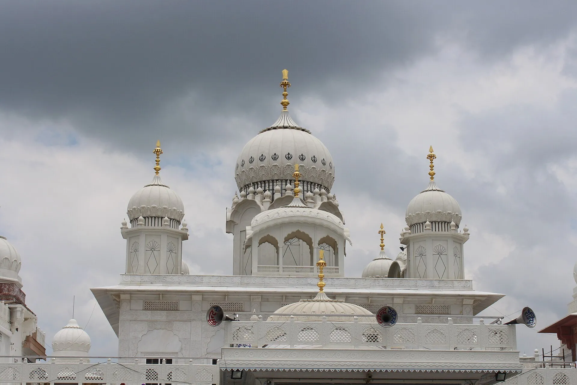 Cúpulas brancas, pináculos dourados e fachada trabalhada do gurdwara Guru ka Tal, sob nuvens.