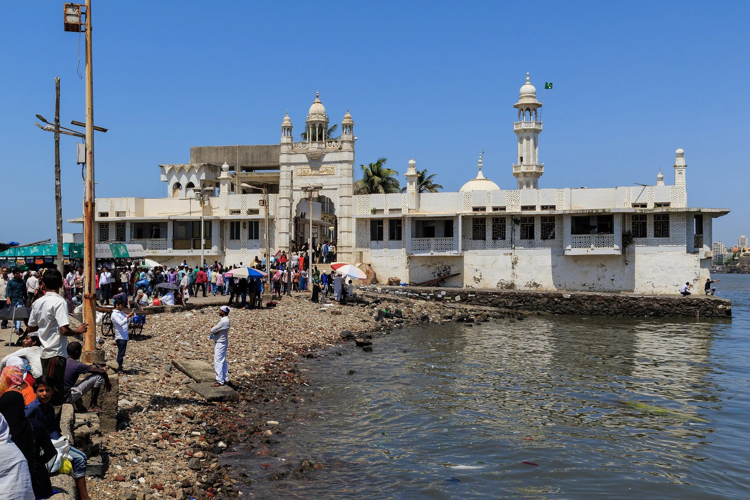 Dargah branco de Haji Ali, em Mumbai, com minarete e cúpulas à beira da água, sob céu azul, e visitantes na margem.
