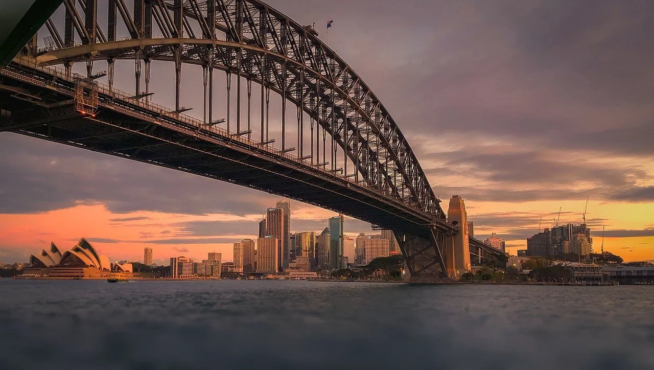 Estrutura da Harbour Bridge sobre a baía, com Ópera à esquerda e edifícios ao fundo sob céu de fim de tarde.