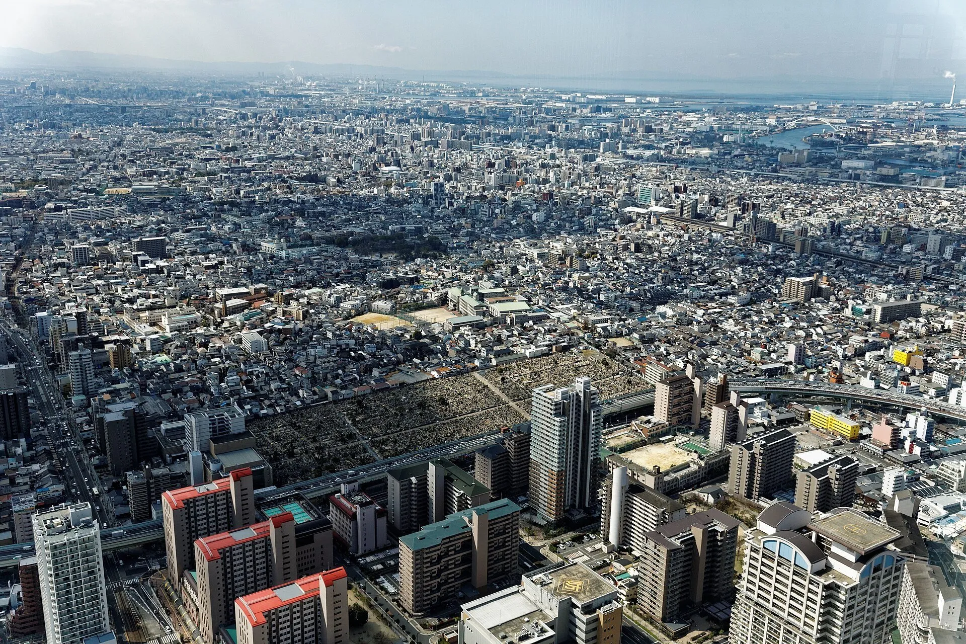 Vista diurna de Osaka a partir do Harukas 300, com edifícios, avenidas e água ao fundo.