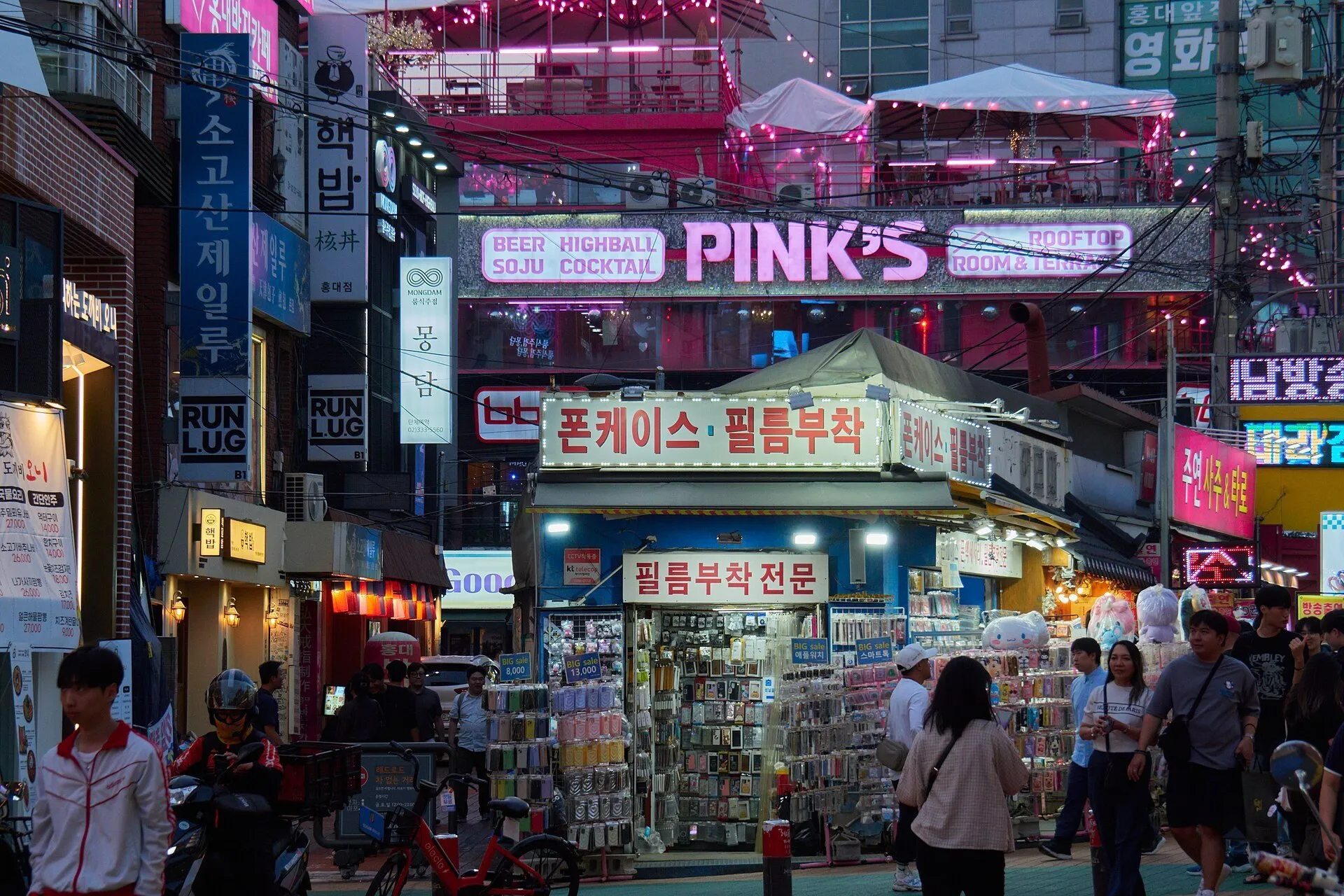 Lojas e letreiros iluminados em uma rua de Hongdae, com pedestres e uma banca de acessórios.