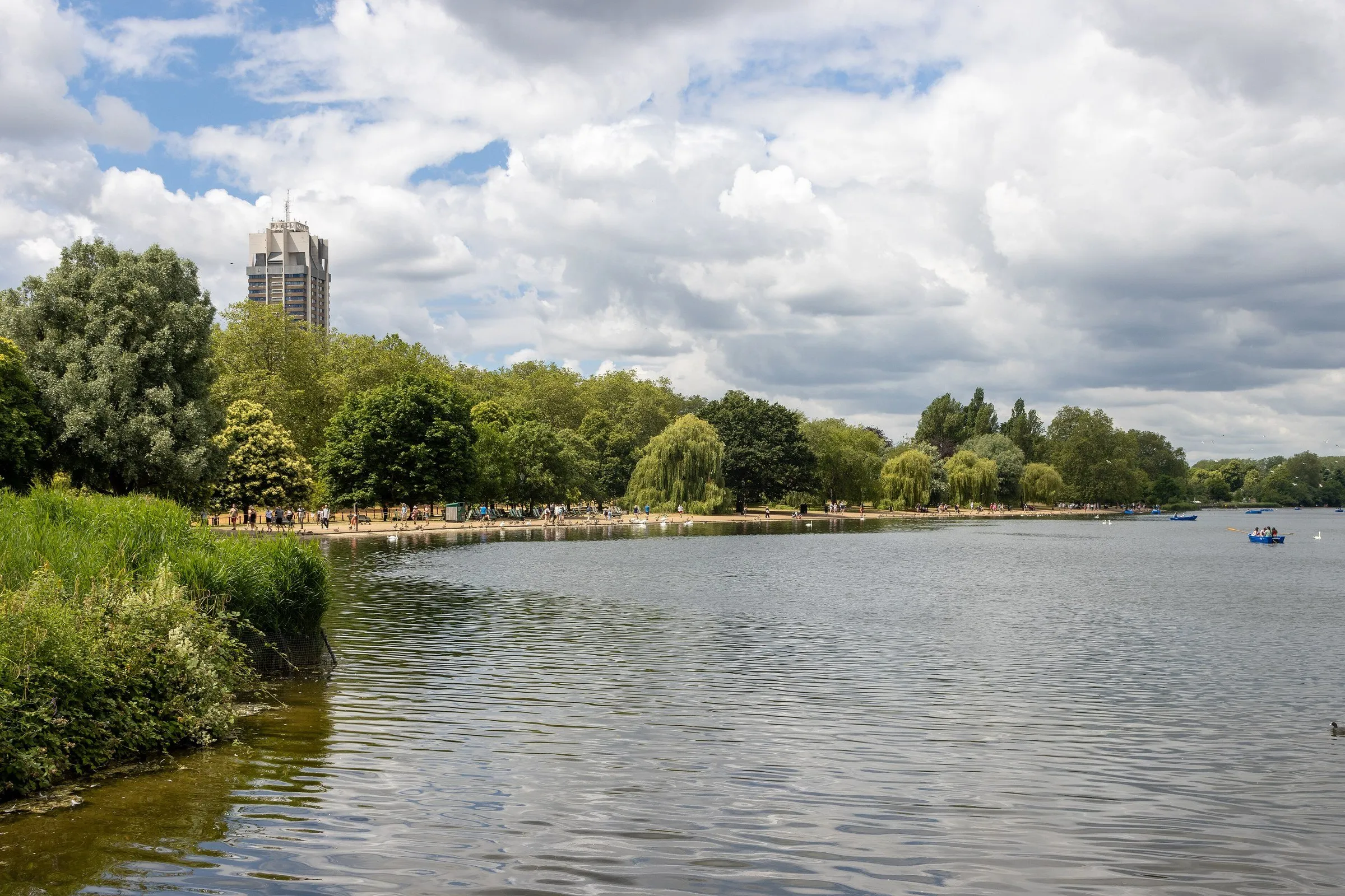 Lago Serpentine no Hyde Park, com árvores à margem, frequentadores na orla e barcos a remo ao longe sob céu nublado.