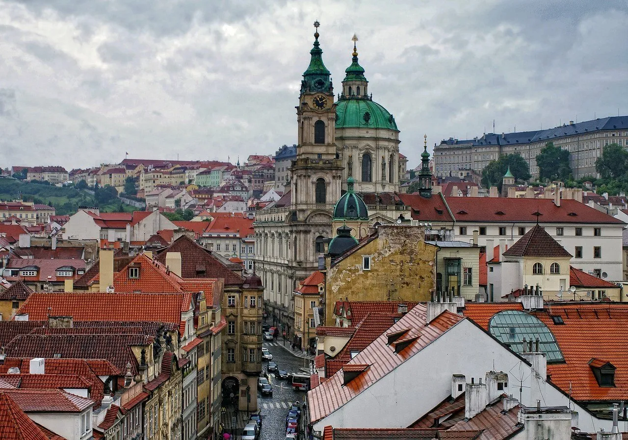 Torre e cúpula verde da Igreja de São Nicolau de Malá Strana, entre telhados vermelhos sob céu nublado.