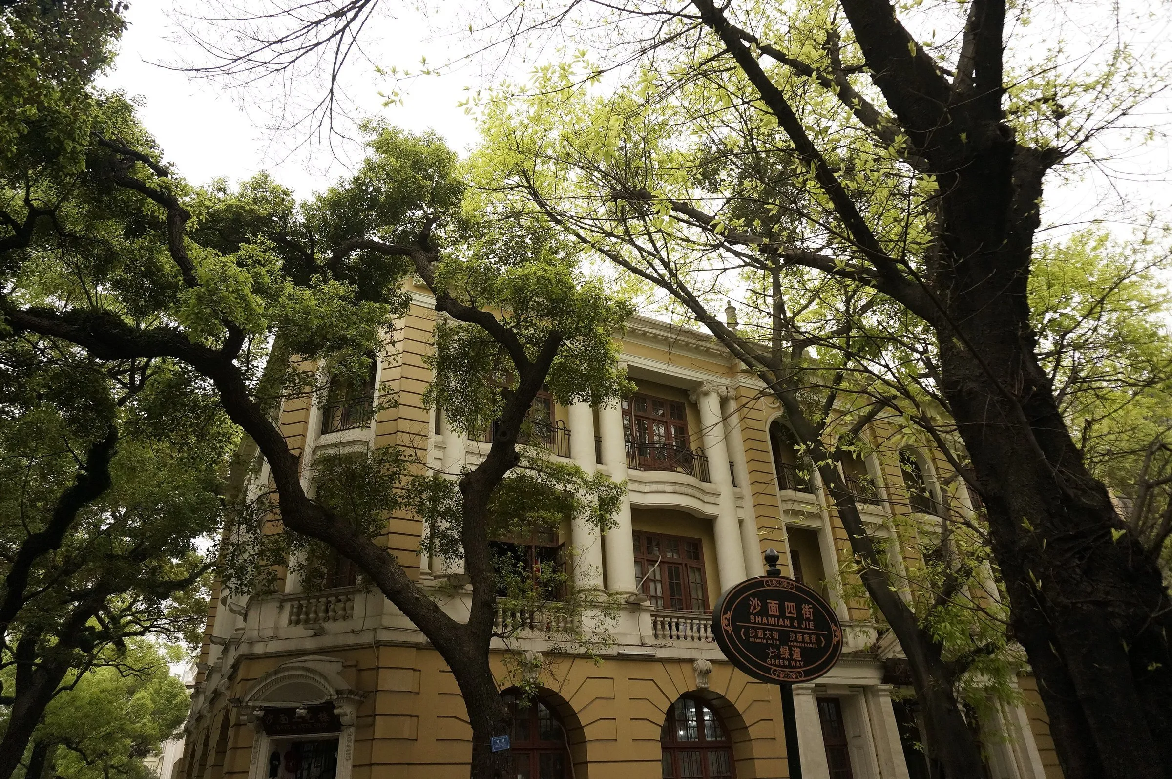 Fachada amarela com colunas e varandas entre árvores na ilha de Shamian, com placa da rua em primeiro plano.