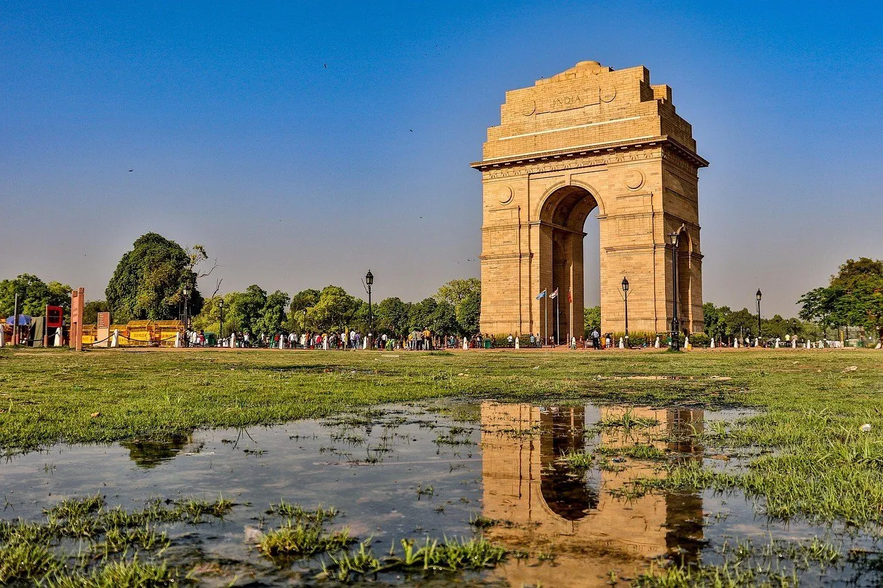 India Gate, em Nova Délhi, de arenito claro sob céu azul, com seu reflexo numa poça no gramado em primeiro plano.