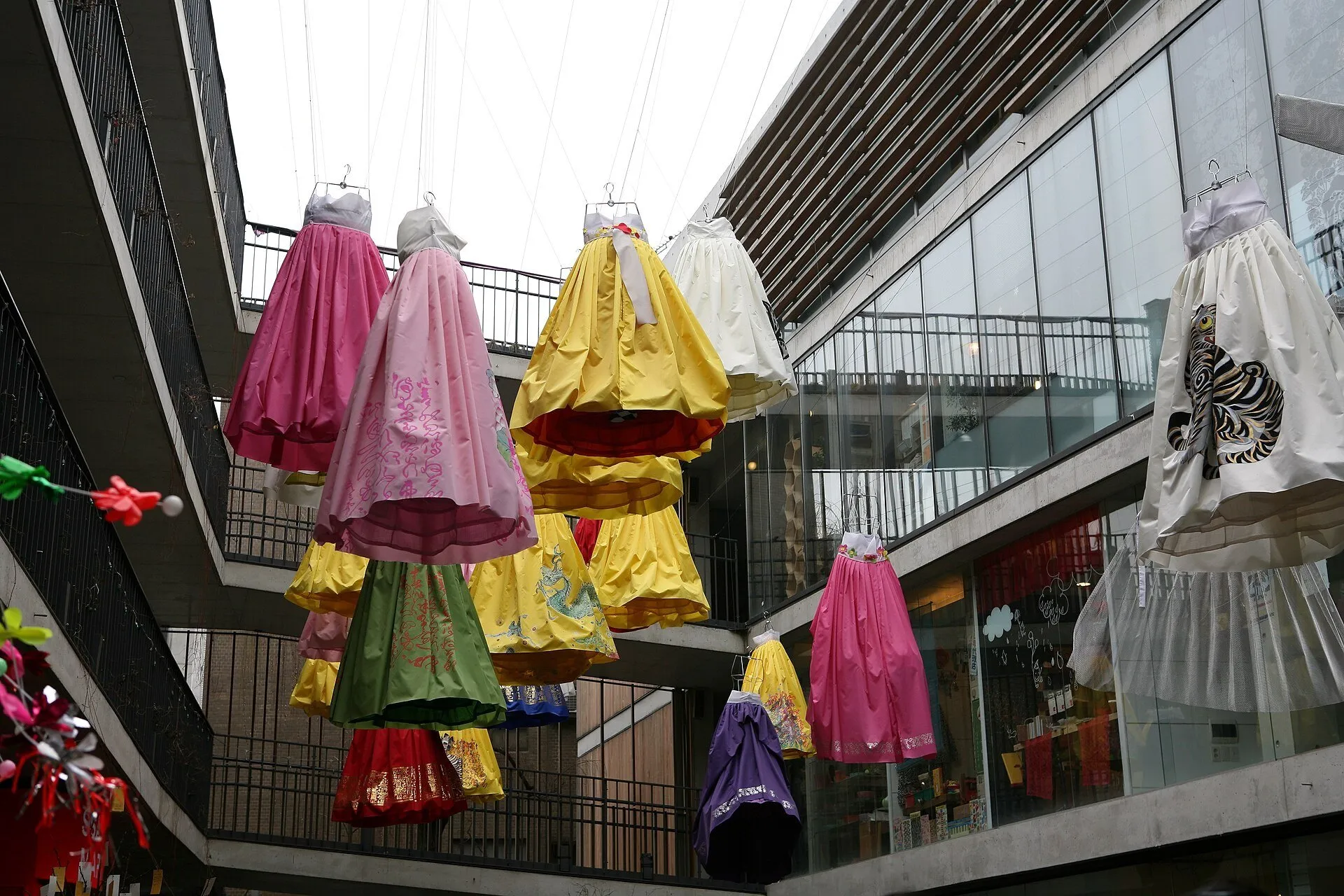 Vestidos coloridos suspensos sobre um pátio comercial em Insadong, com lojas e passarelas ao redor.