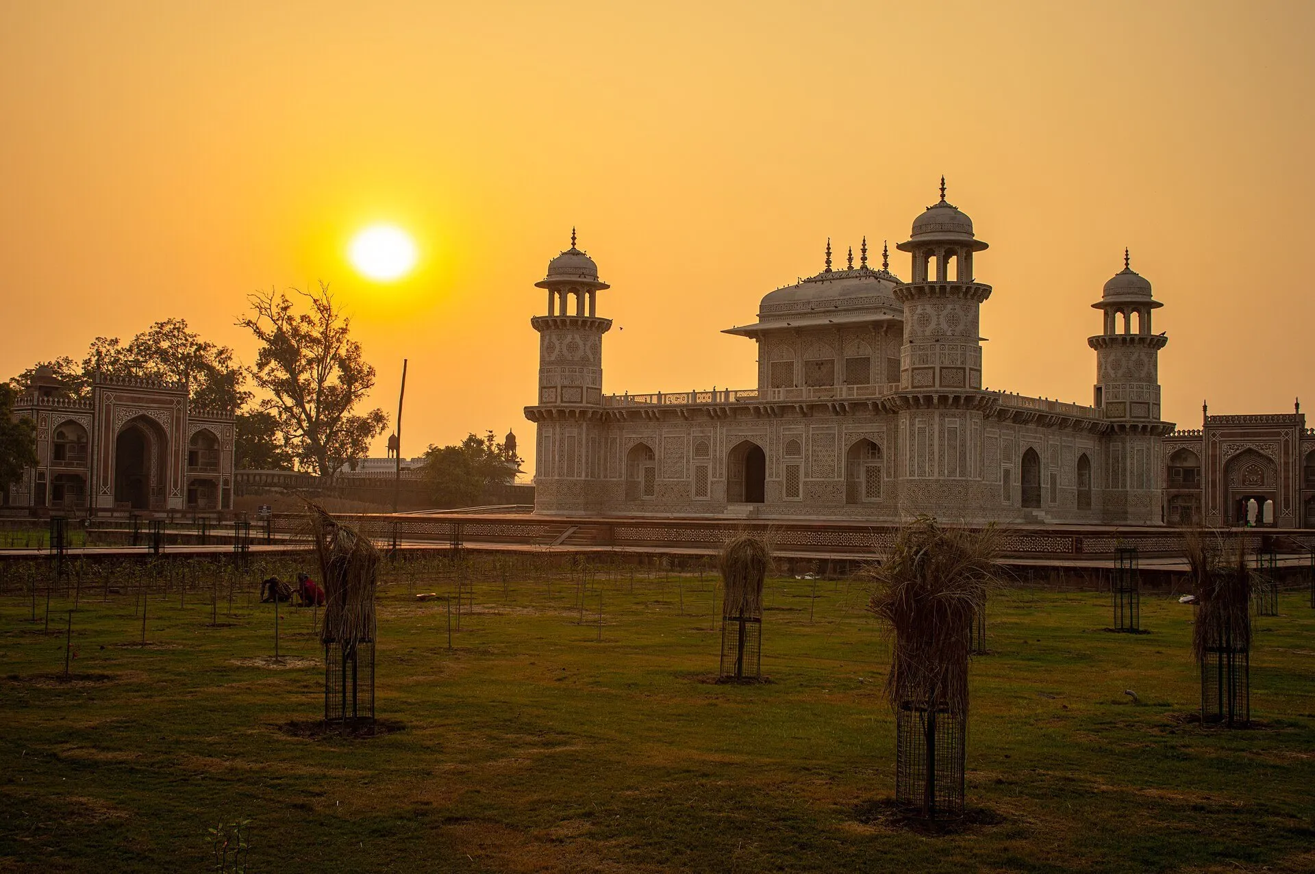Tumba de Itimad-ud-Daulah e suas torres de canto diante do sol baixo, com jardim no primeiro plano.