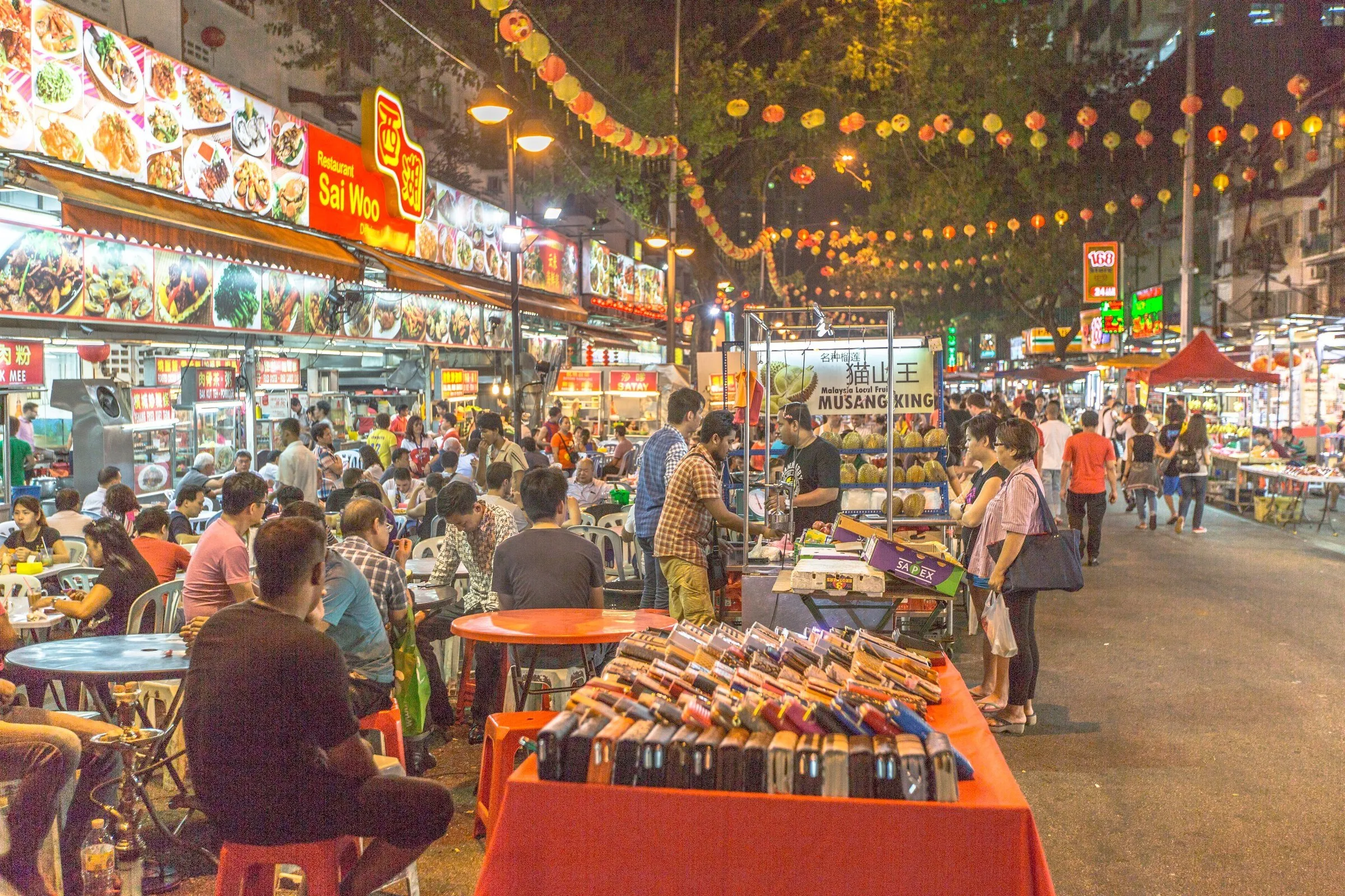 Rua de comida à noite, com mesas cheias na calçada, painéis de pratos, banca de duriana e lanternas acesas penduradas.
