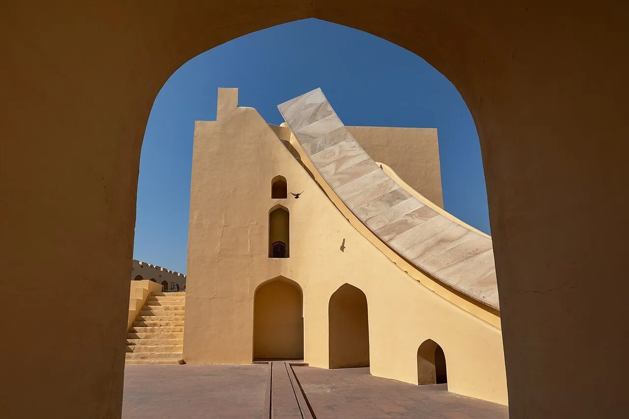 Instrumento de alvenaria clara do Jantar Mantar de Jaipur, enquadrado por um arco com céu azul ao fundo.