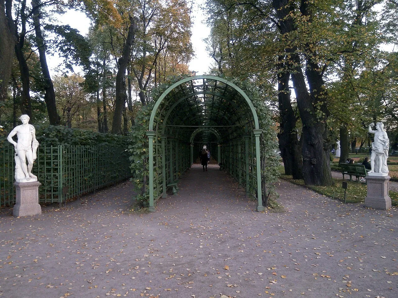 Pérgola verde ladeada por esculturas brancas e árvores no Jardim de Verão, com folhas no chão.