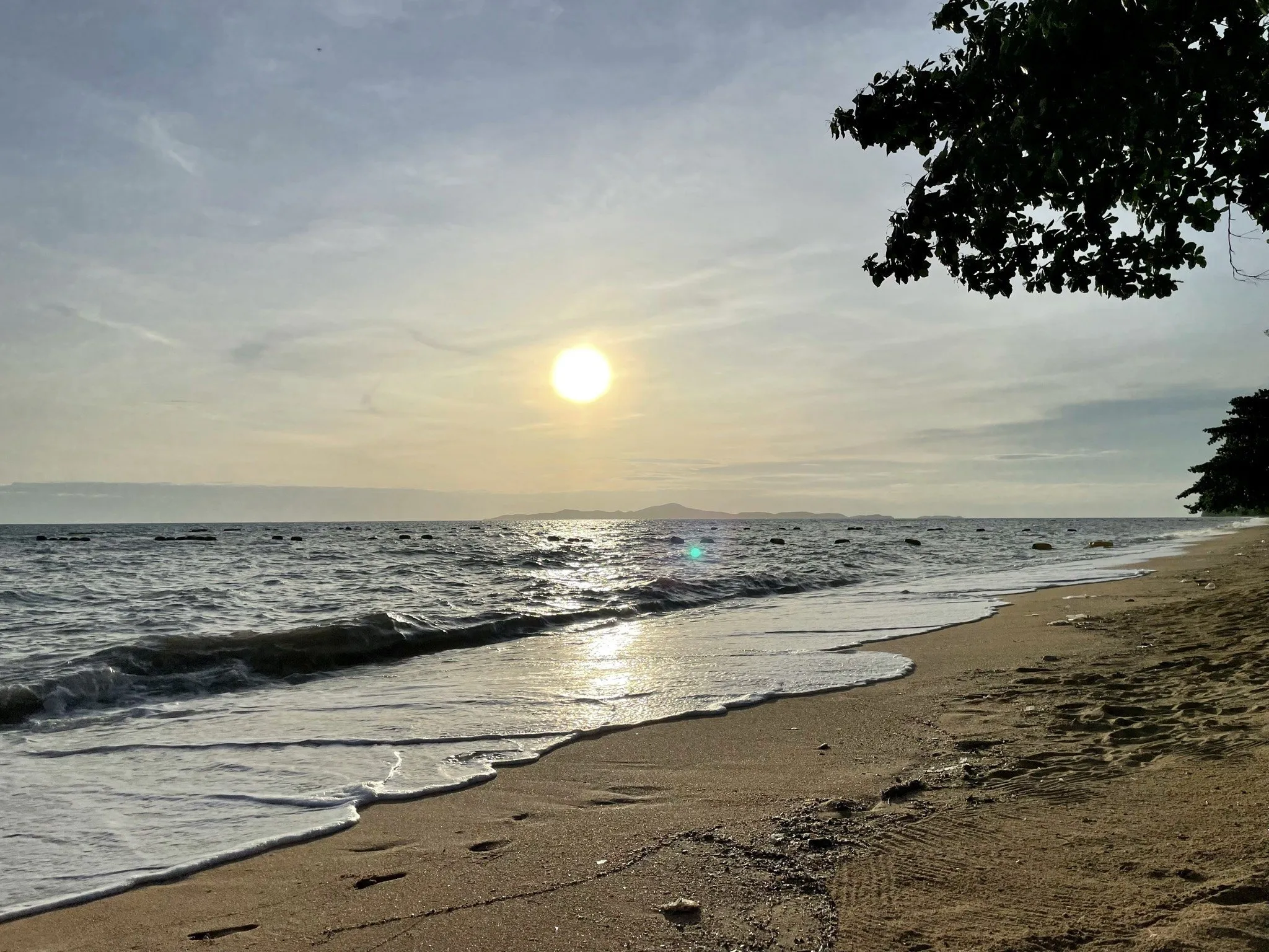 Praia de Jomtien ao entardecer, com sol refletido no mar, ondas na areia e galhos no alto do quadro.