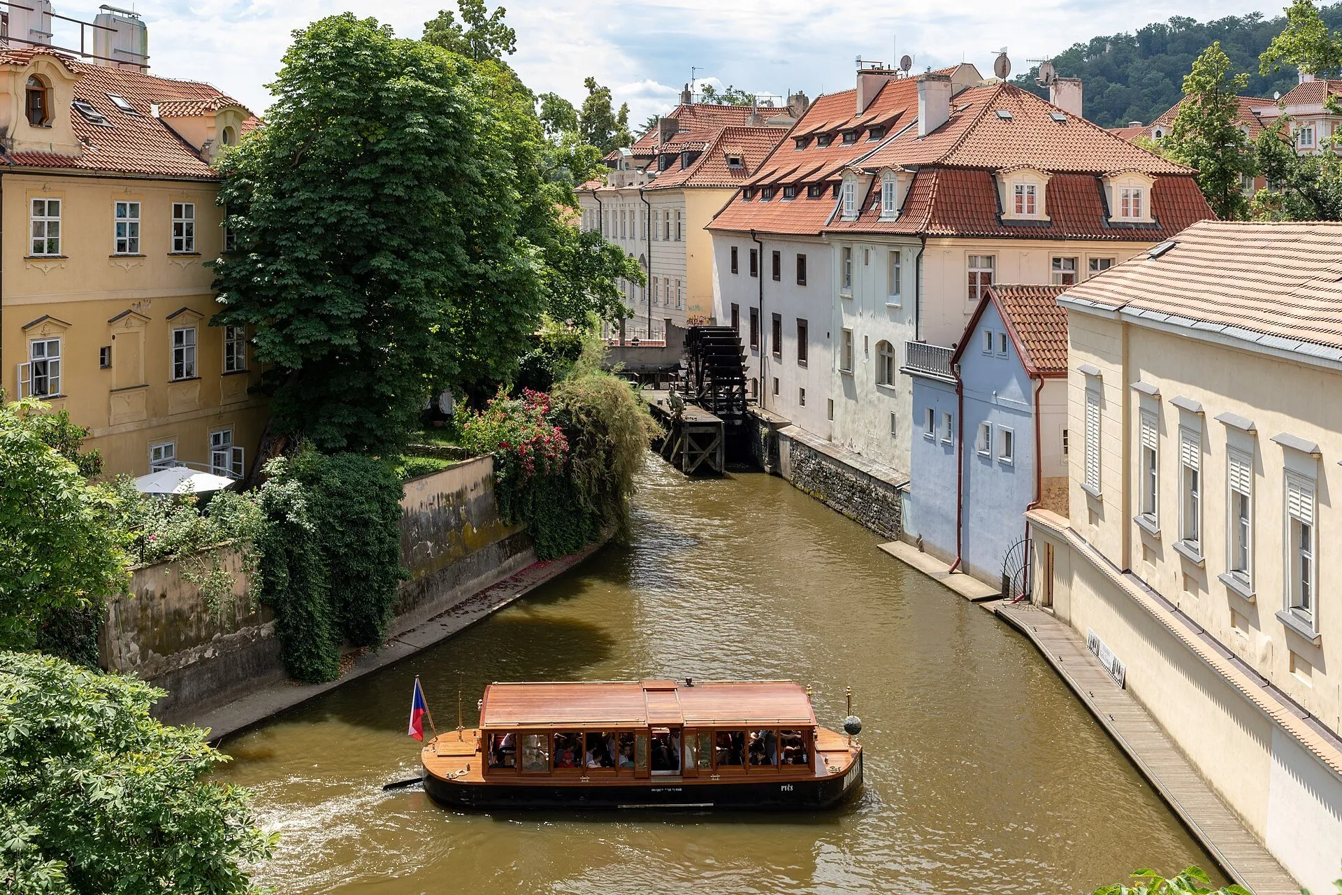 Canal Čertovka em Kampa, com roda de moinho, casas de telhados vermelhos, árvores e um barco de passeio.