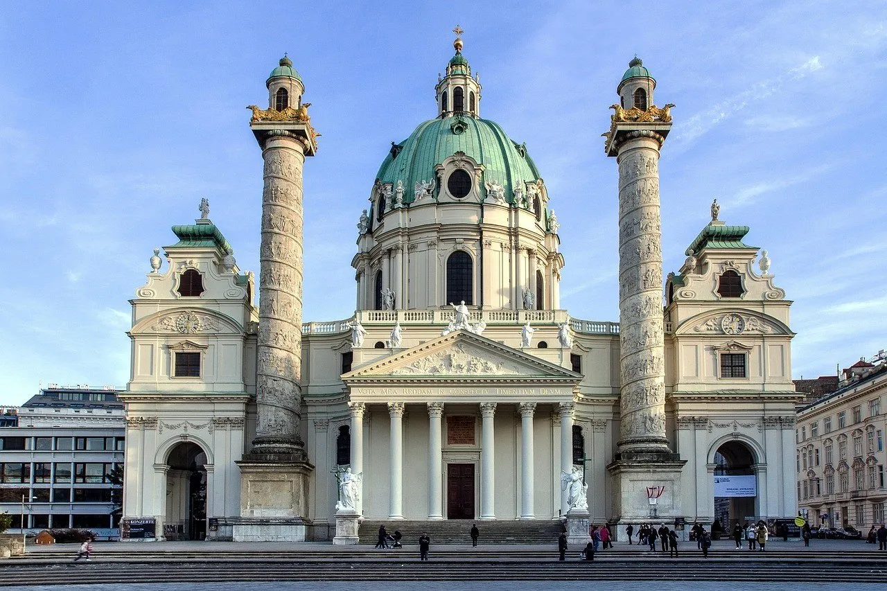 Fachada da Karlskirche com cúpula verde e duas colunas, diante de escadaria com visitantes.