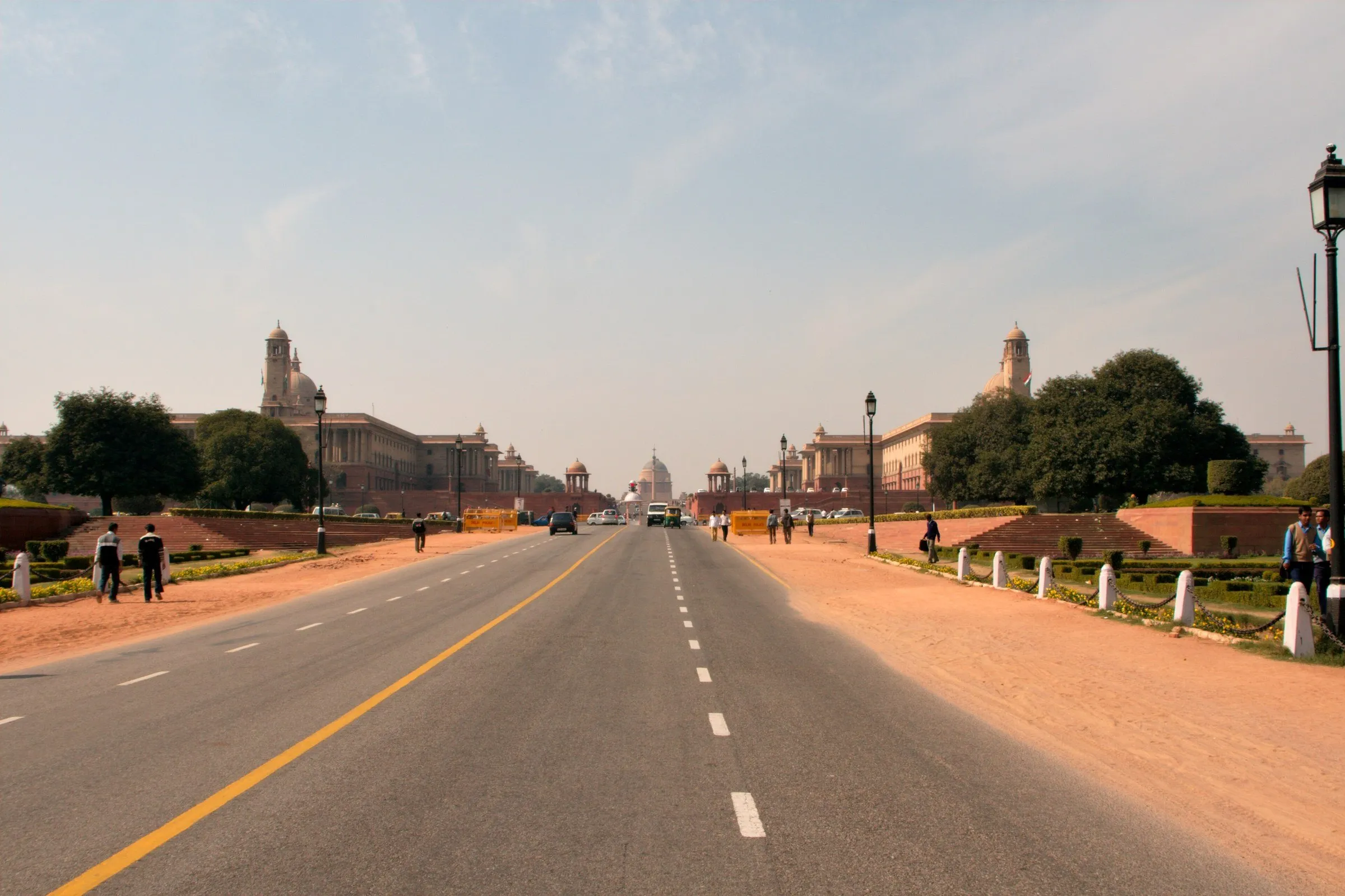 Avenida larga e reta com os prédios do Secretariado dos dois lados e a cúpula do Rashtrapati Bhavan ao fundo, sob céu claro.