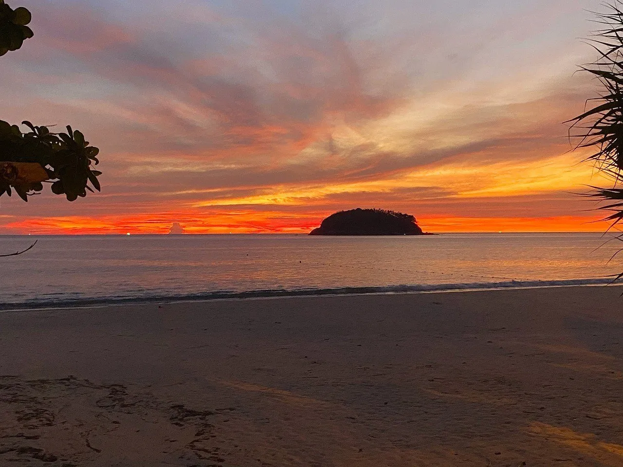 Praia de Kata, em Phuket, ao pôr do sol, com areia clara em primeiro plano e a ilhota Koh Pu recortada contra o céu laranja.