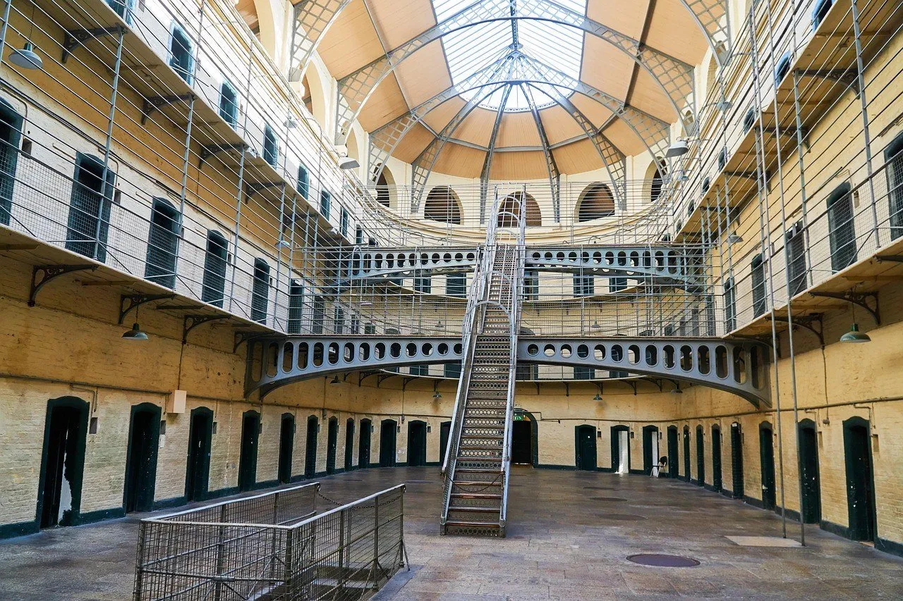 Salão de Kilmainham Gaol com escadaria central, passarelas metálicas, portas de celas e teto envidraçado.