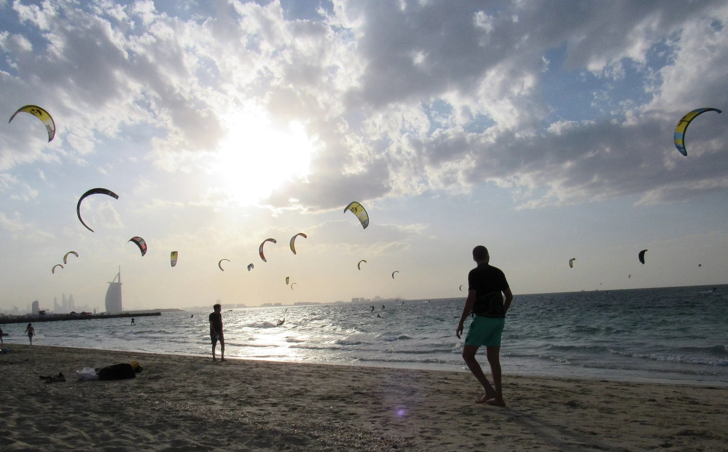 Dezenas de kites no céu nublado sobre o mar em Kite Beach, com a silhueta do Burj Al Arab ao fundo e banhistas na areia.