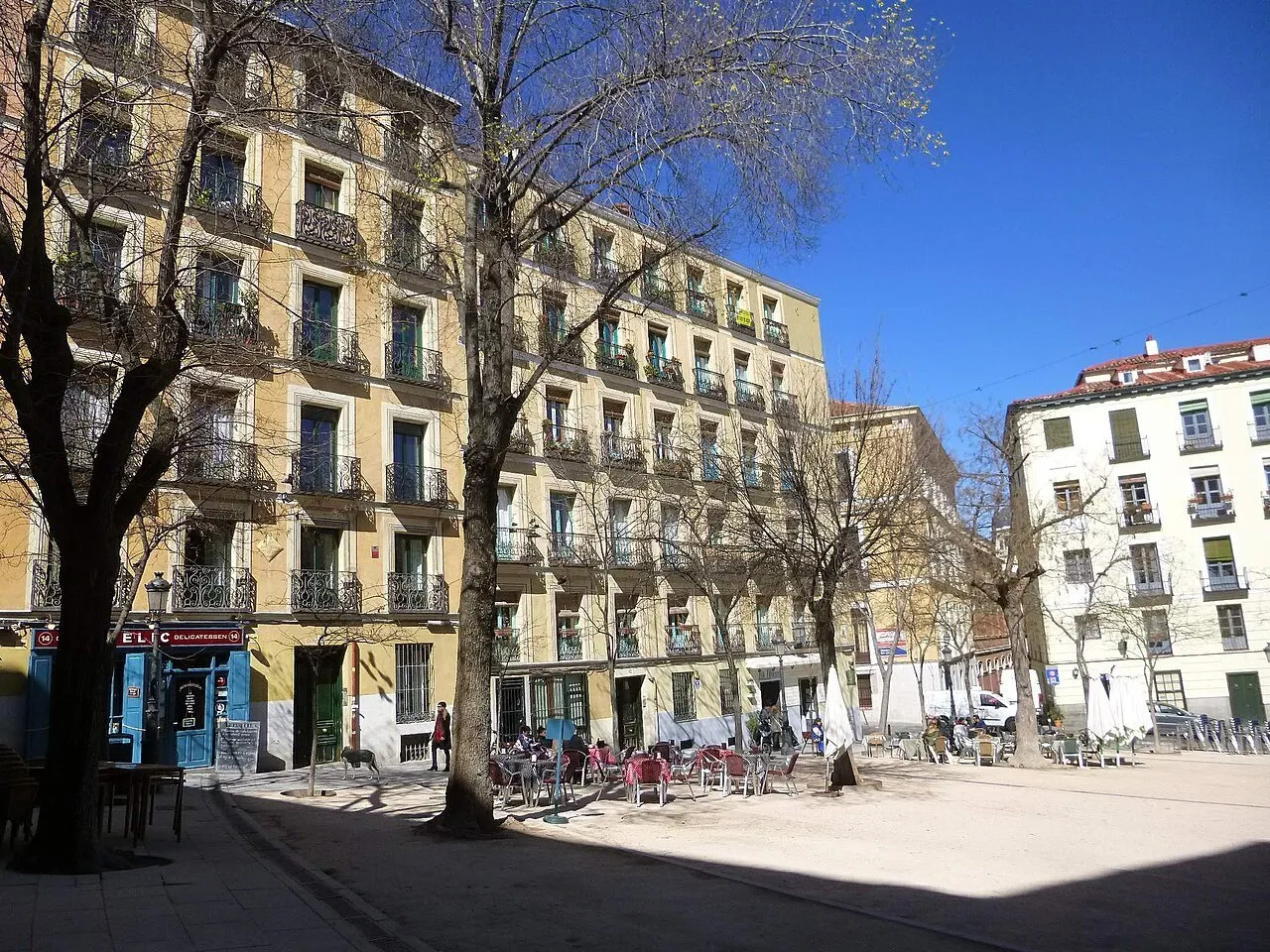 Plaza de la Paja, em La Latina, com árvores, fachadas amarelas e brancas e mesas ao ar livre.