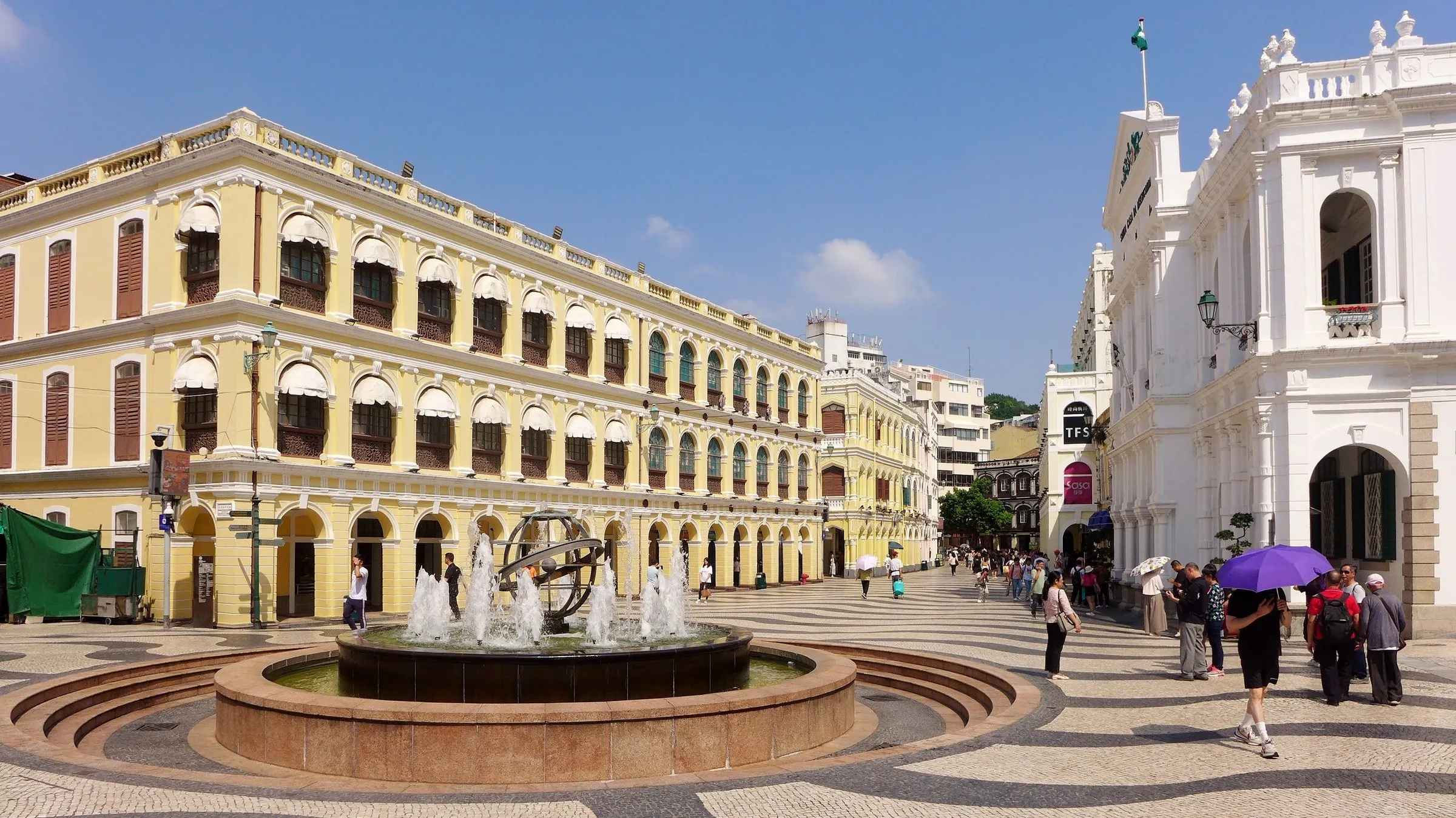 Praça com calçada portuguesa em ondas preto e branco, fonte ao centro e prédios históricos amarelos e brancos sob céu azul.