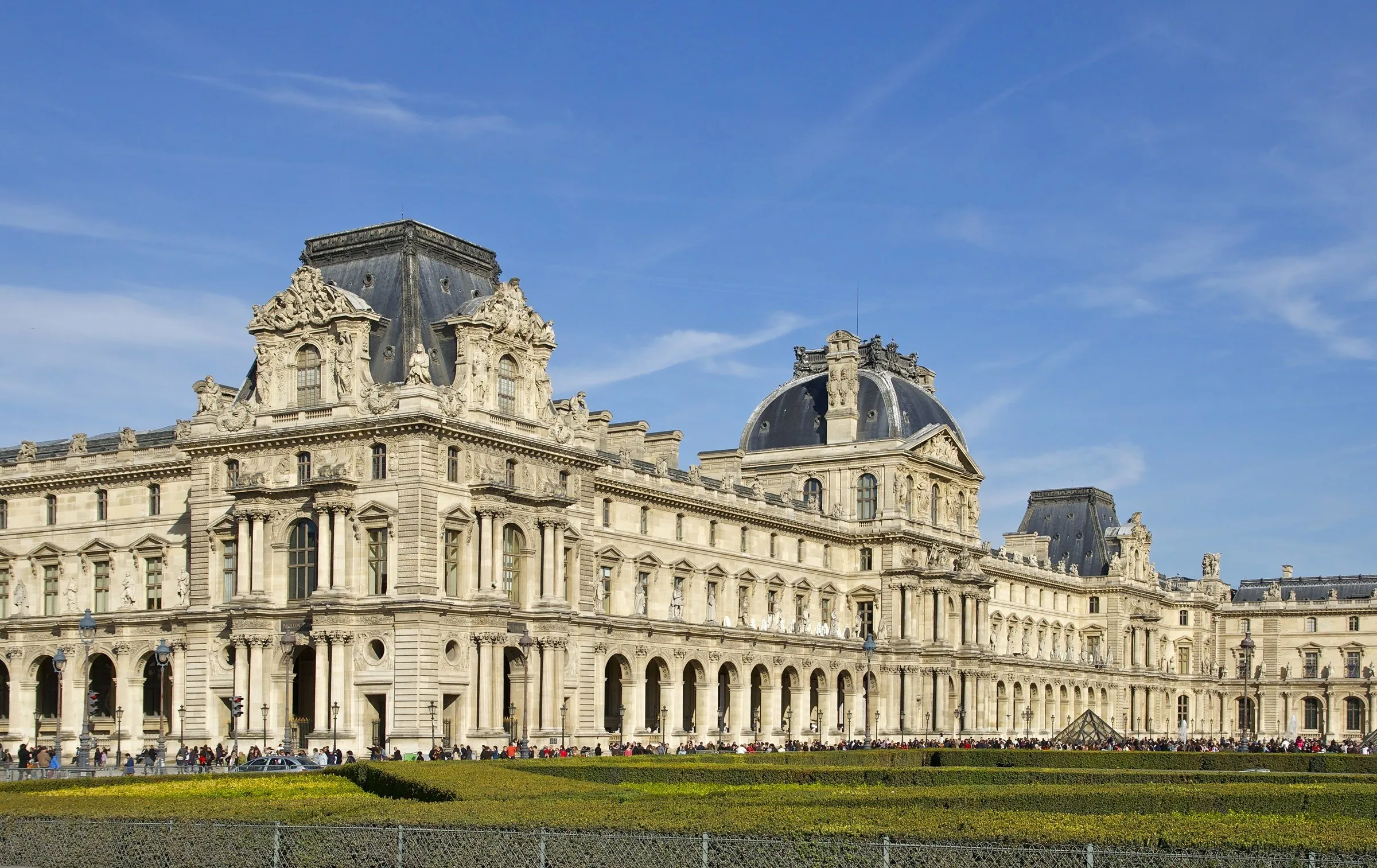 Fachada da ala Richelieu do Palácio do Louvre, em pedra clara, com telhados de ardósia sob céu azul com poucas nuvens.