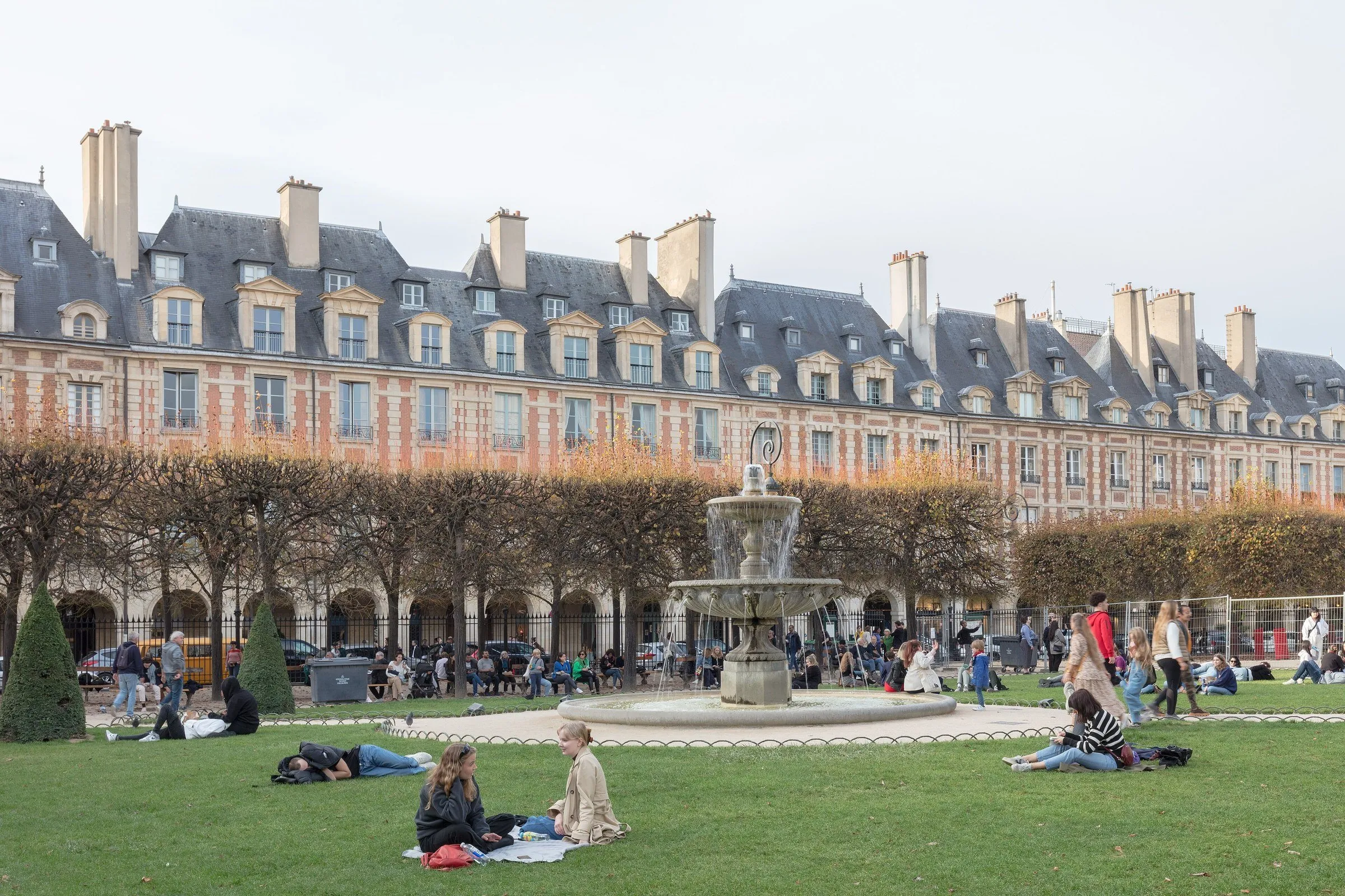 Fonte e gramado da Place des Vosges, em Paris, com pessoas na grama e casas de tijolo com telhado de ardósia ao fundo.