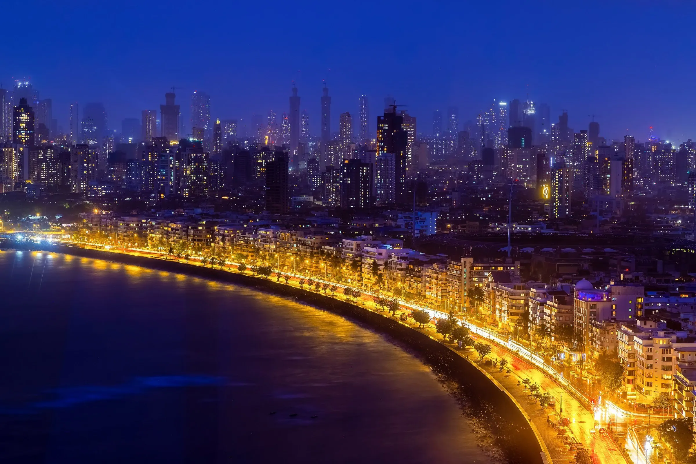 Vista aérea noturna da curva da Marine Drive, em Mumbai, com a avenida iluminada à beira-mar e arranha-céus ao fundo.