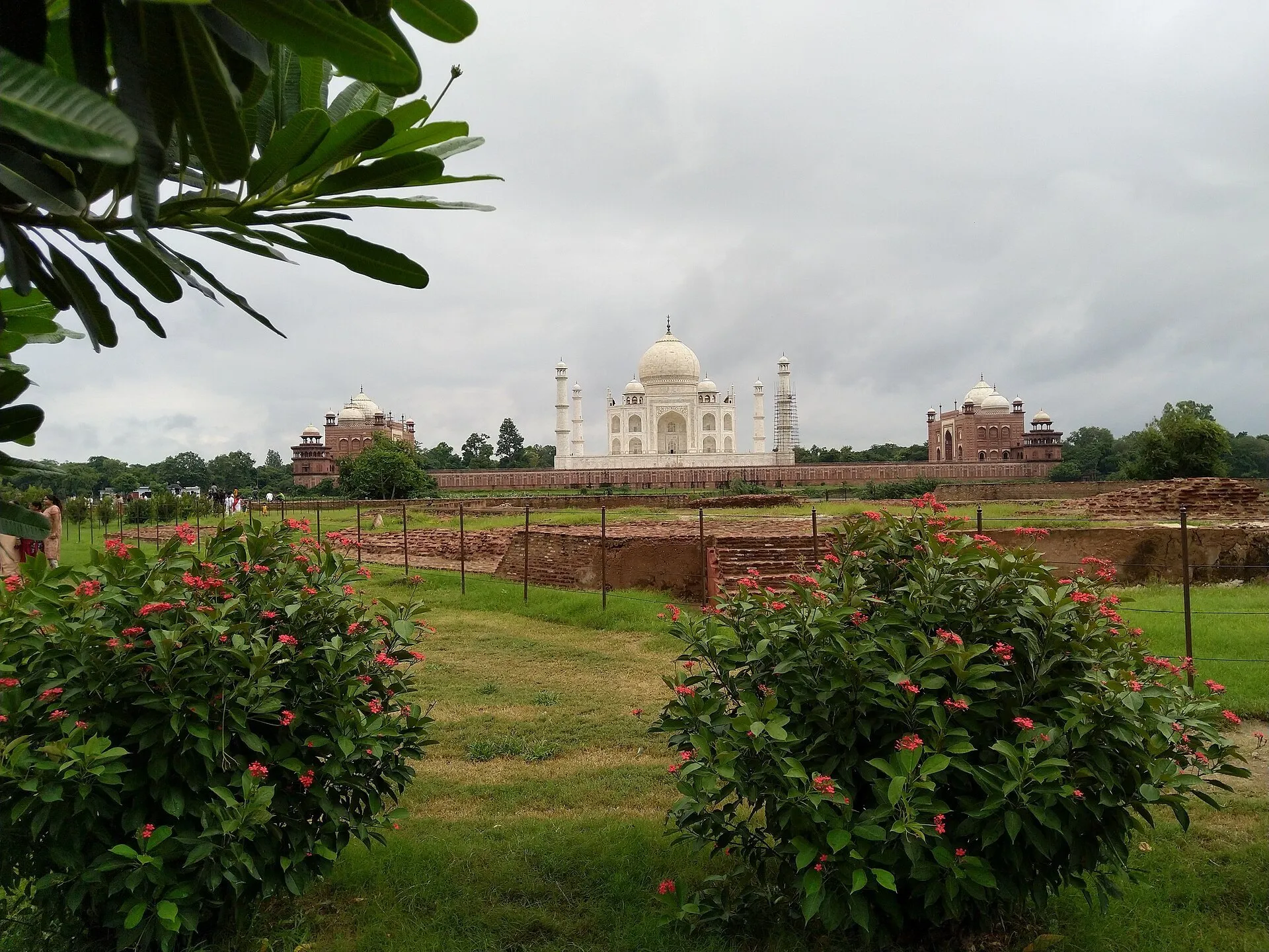 Jardim de Mehtab Bagh com flores e ruínas baixas; Taj Mahal ao fundo, com um minarete em restauro.