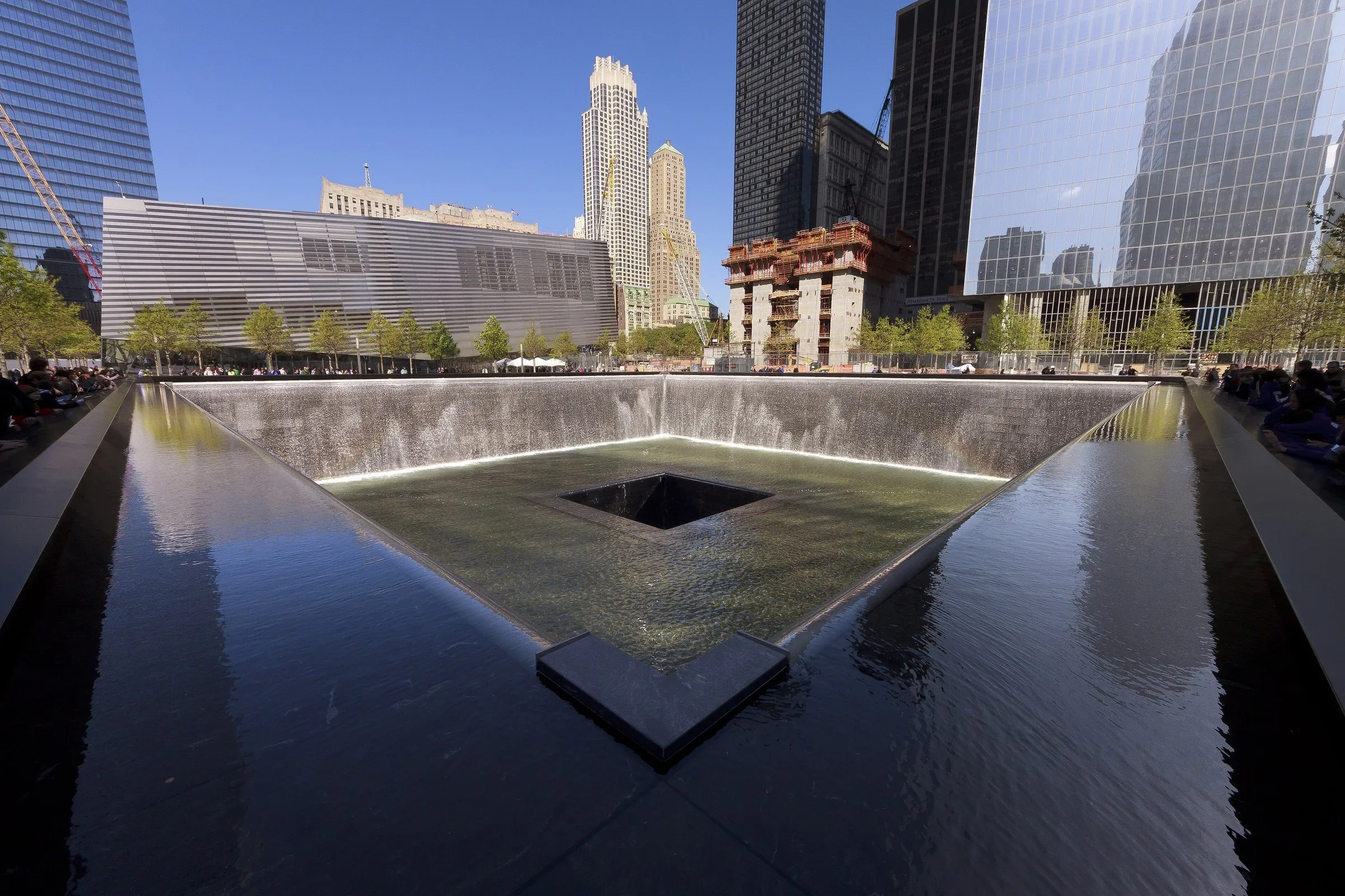 Espelho d'água quadrado do Memorial do 11 de Setembro, com cascata nas bordas, bronze em primeiro plano e prédios ao redor.