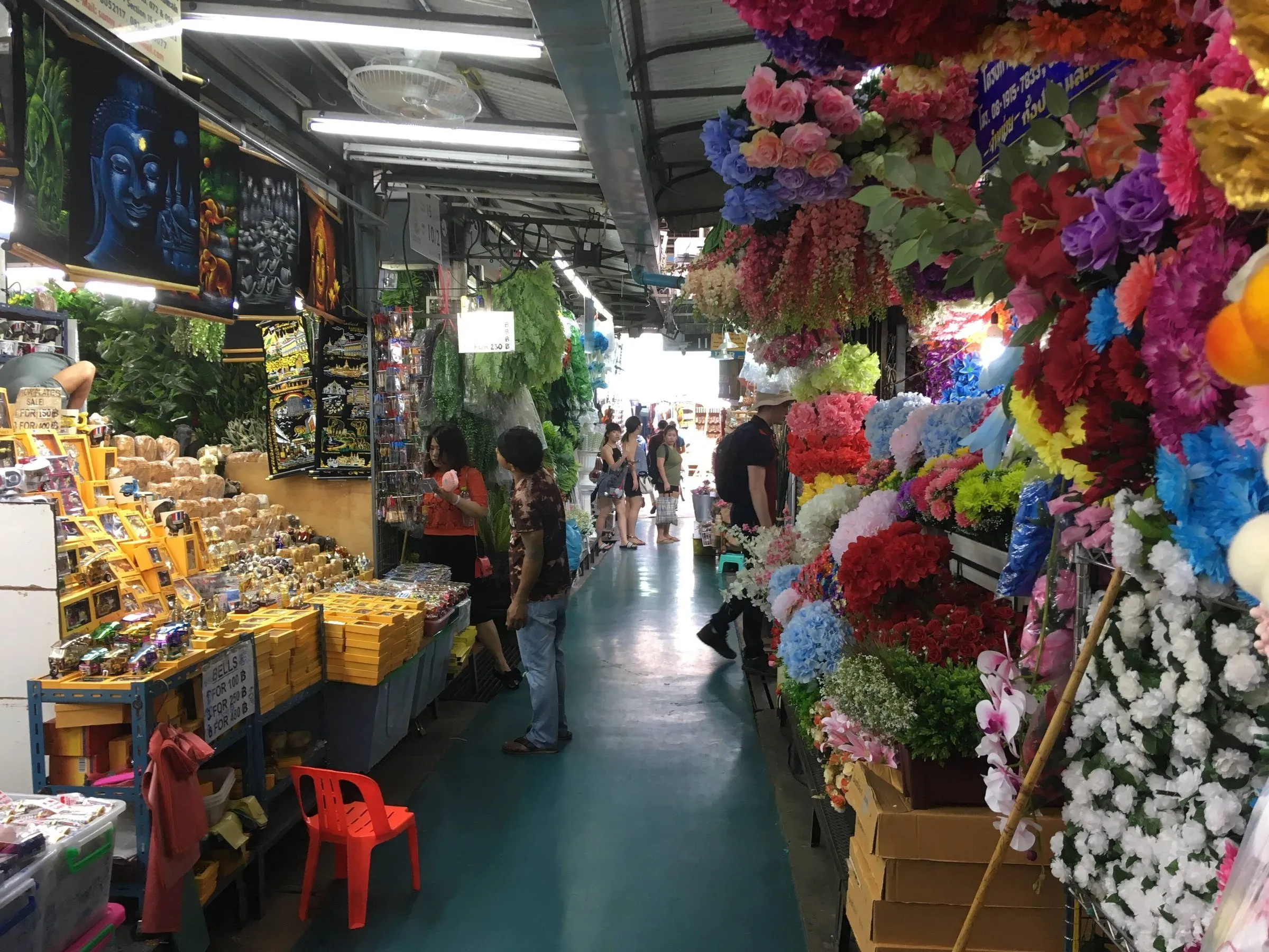 Corredor coberto do mercado Chatuchak, em Bangkok, com lembranças à esquerda e flores artificiais coloridas à direita.