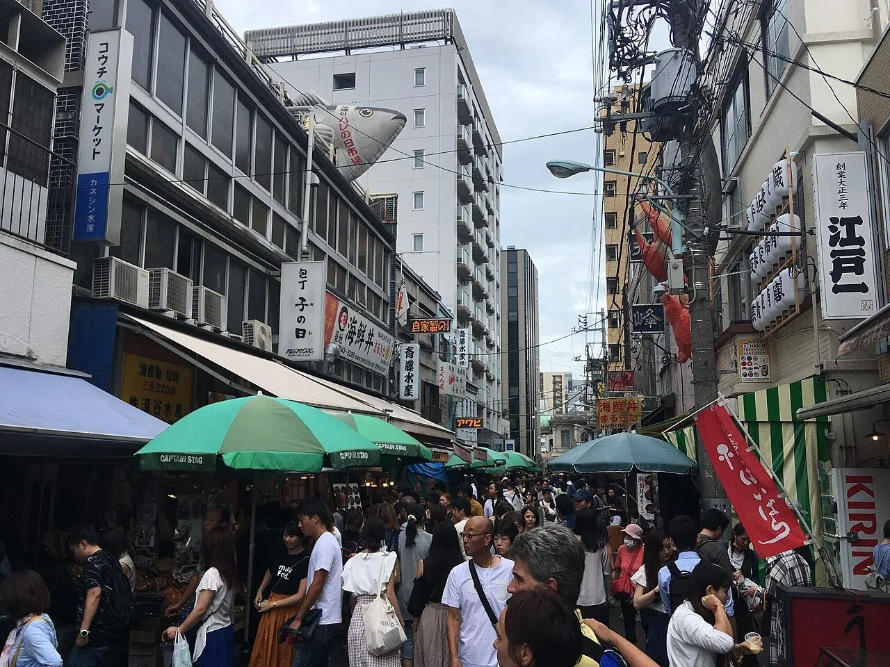 Rua do Mercado Externo de Tsukiji com bancas sob guarda-sóis verdes, letreiros e muitos visitantes.