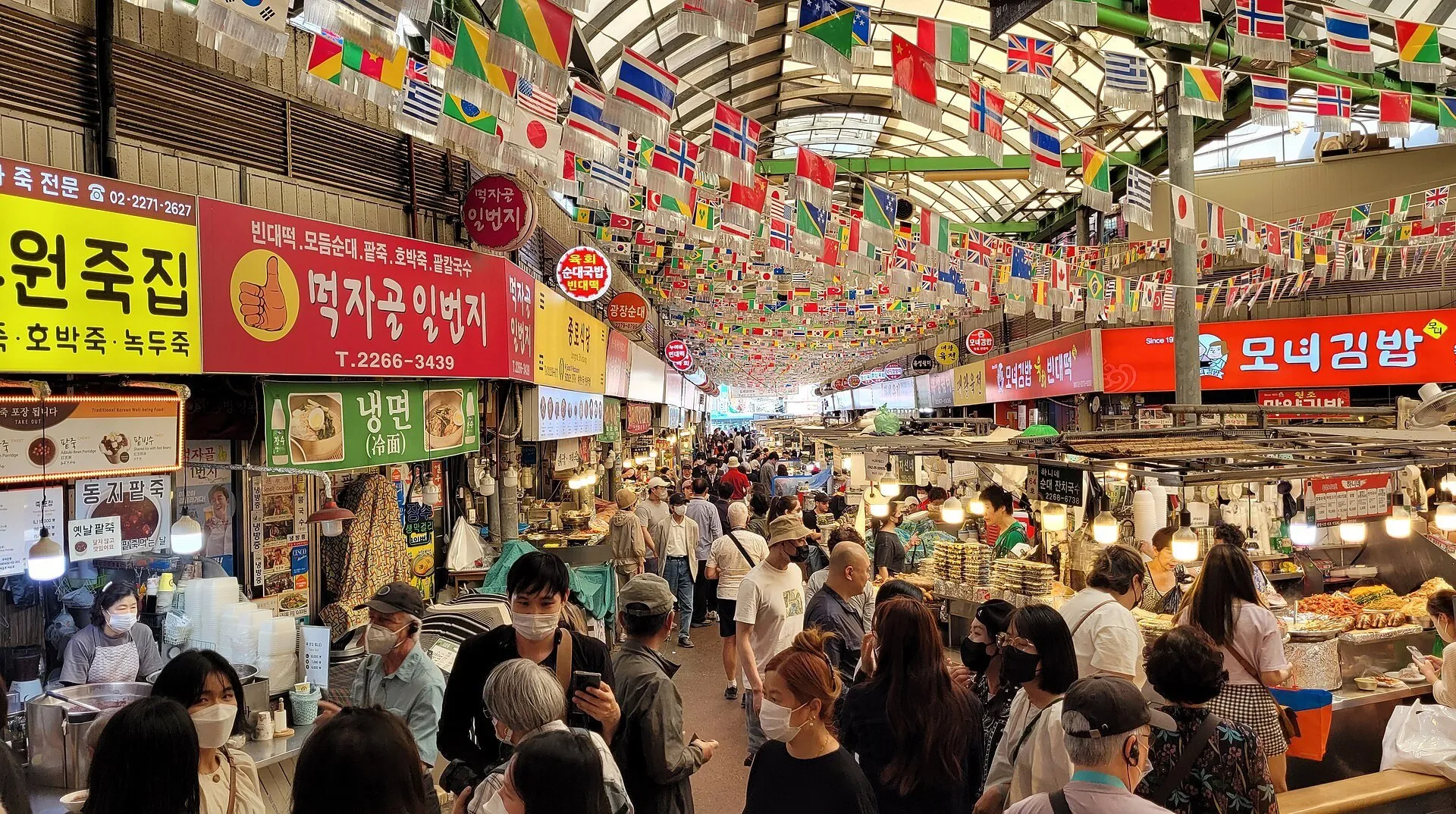 Corredor coberto do Mercado Gwangjang com letreiros, bandeiras, bancas de comida e visitantes.