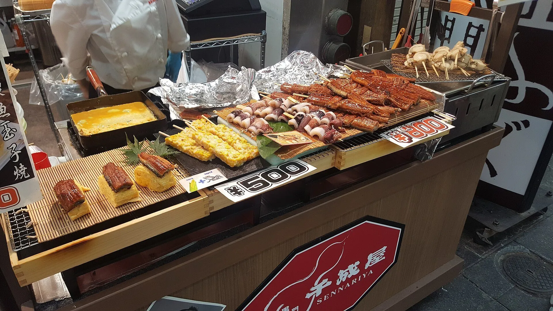 Banca do Mercado Kuromon com porções prontas, espetinhos de frutos do mar e placas de preços.