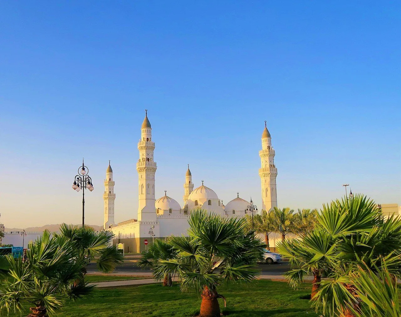 Mesquita de Quba com quatro minaretes claros, cúpulas e palmeiras em primeiro plano, sob luz dourada.