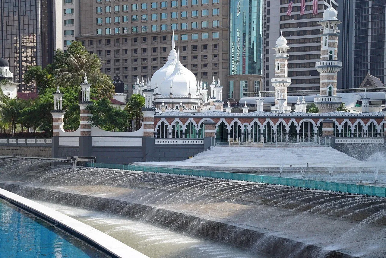 Mesquita Jamek com cúpulas brancas, minaretes de tijolo listrado e arcadas, atrás de um canal com cascata e prédios ao fundo.