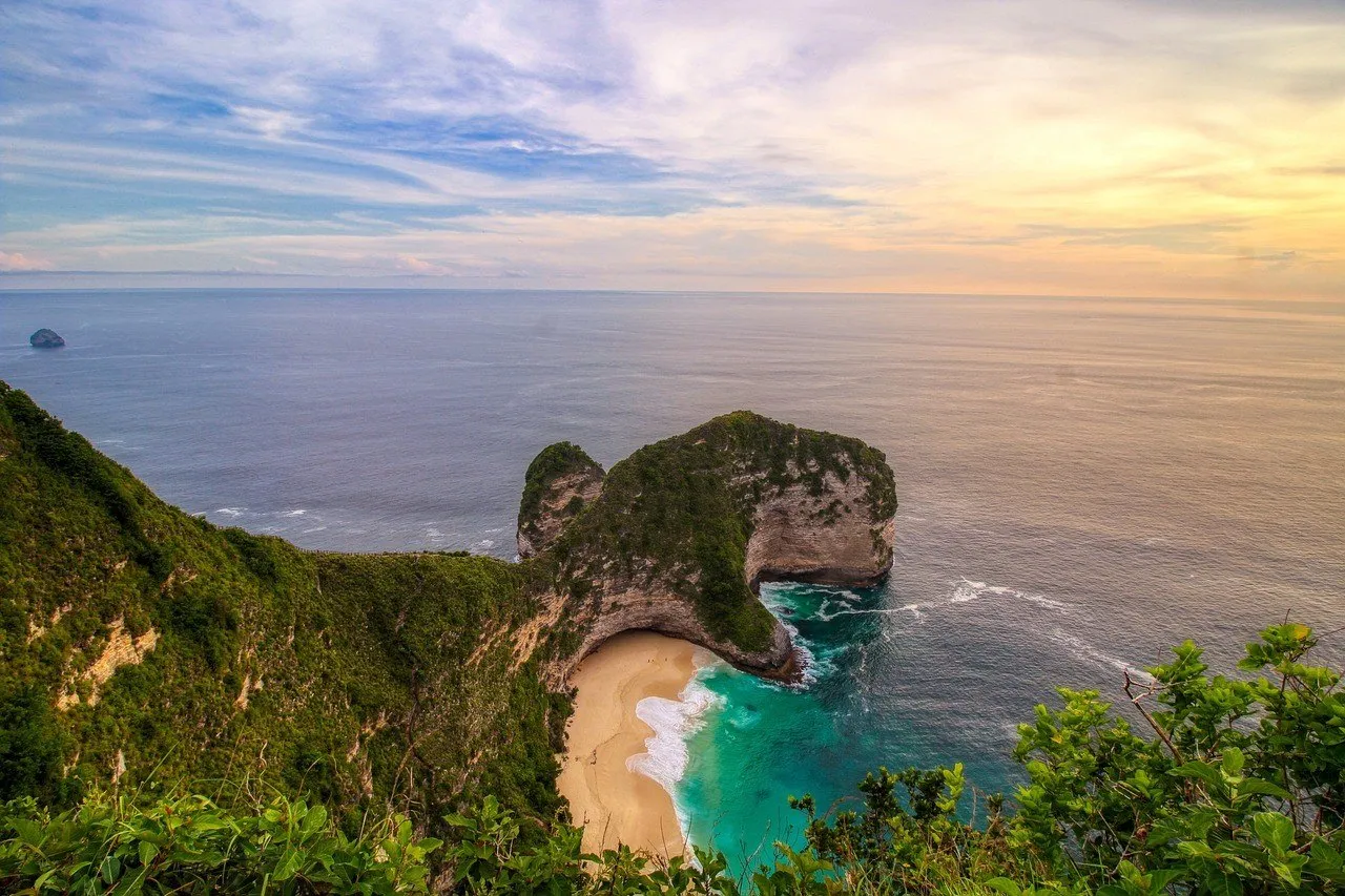 Falésia de Kelingking vista do alto ao entardecer, com praia clara, vegetação e mar azul-esverdeado.