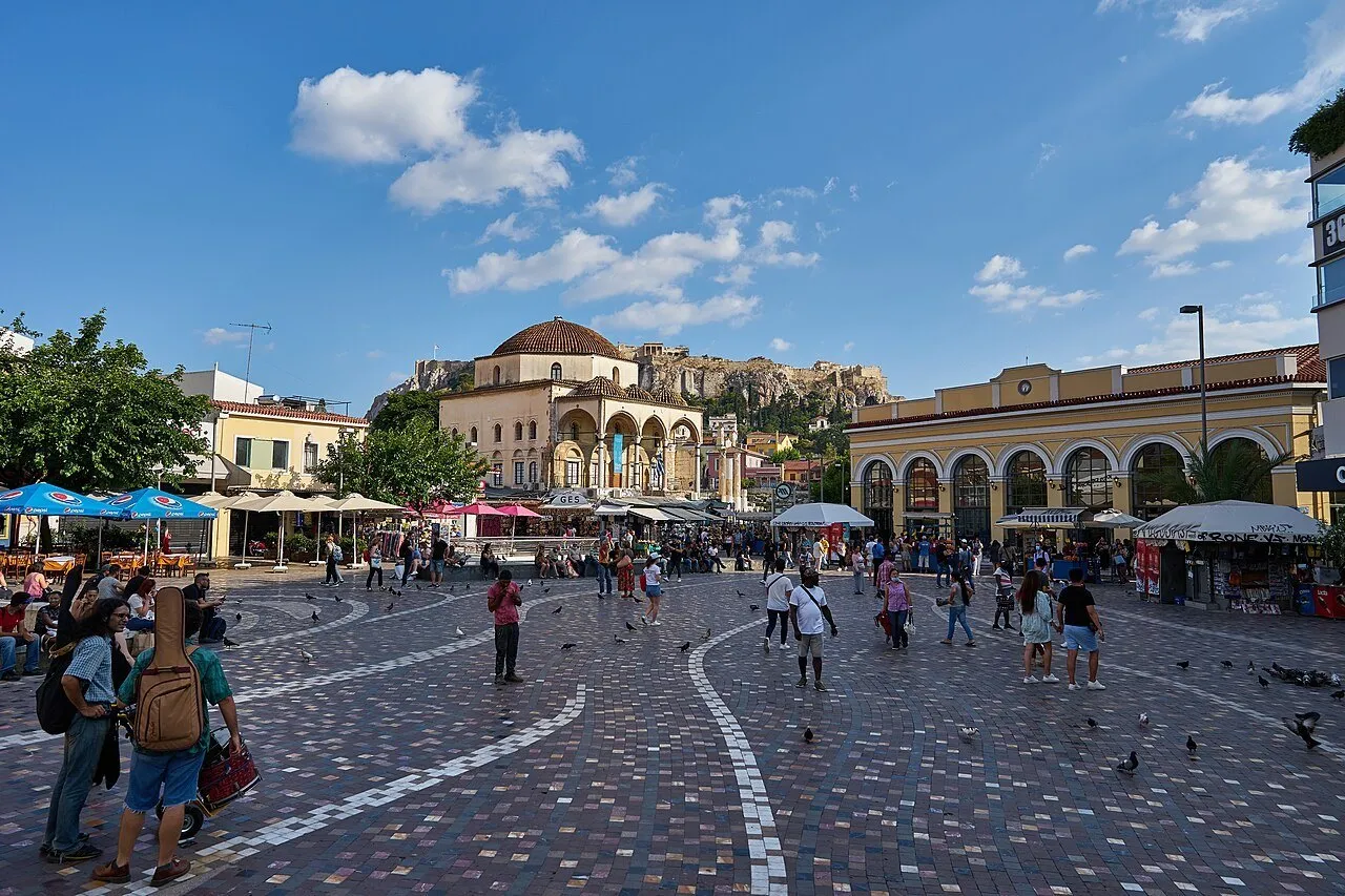 Praça Monastiraki com pedestres, antiga mesquita, estação amarela e Acrópole ao fundo sob céu azul.