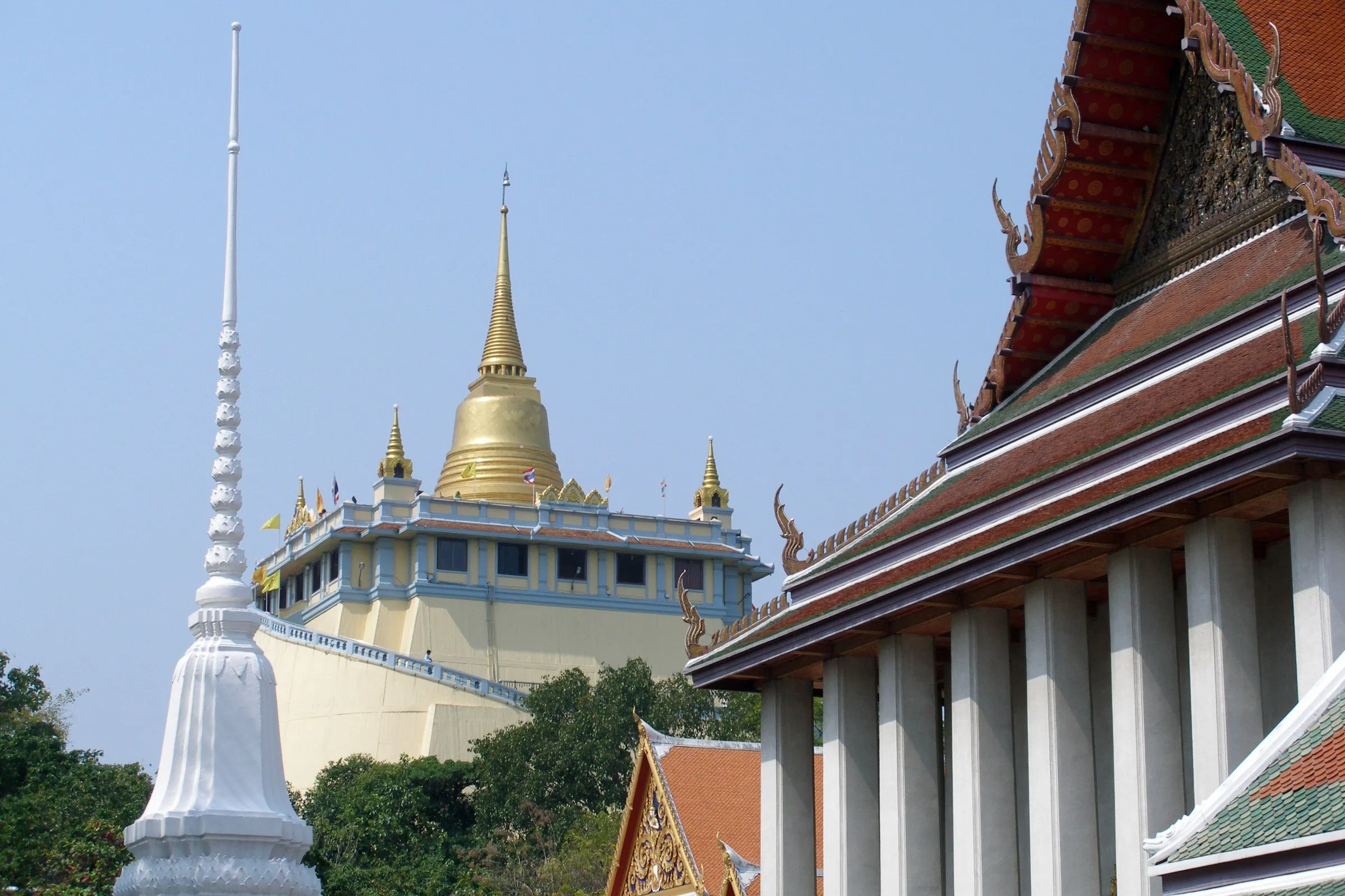 O chedi dourado do Wat Saket (Monte Dourado), em Bangkok, entre um pináculo branco e o beiral de um templo, sob céu azul.